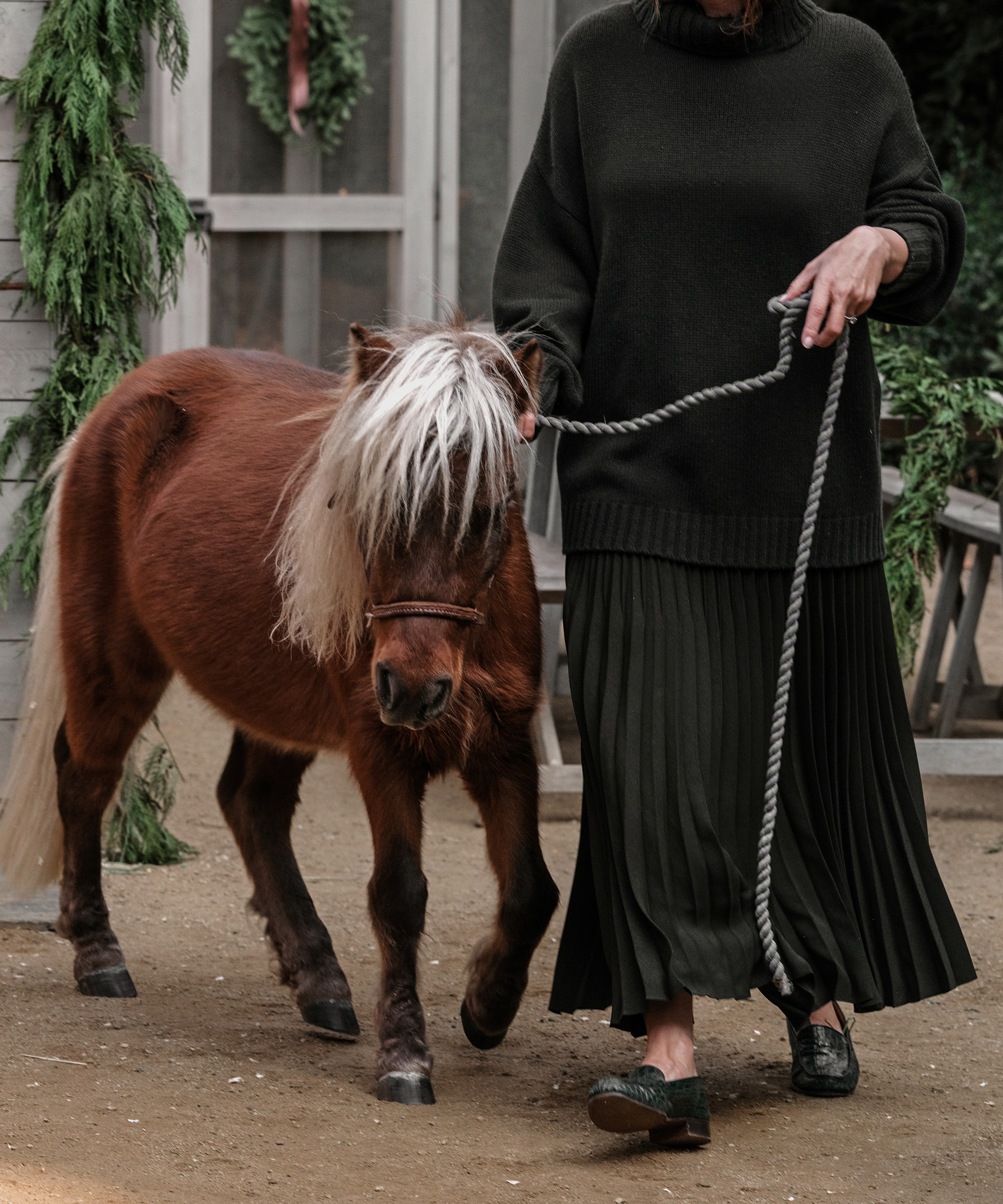 jenni wearing a pleated skirt and a cashmere turtleneck walking with a horse