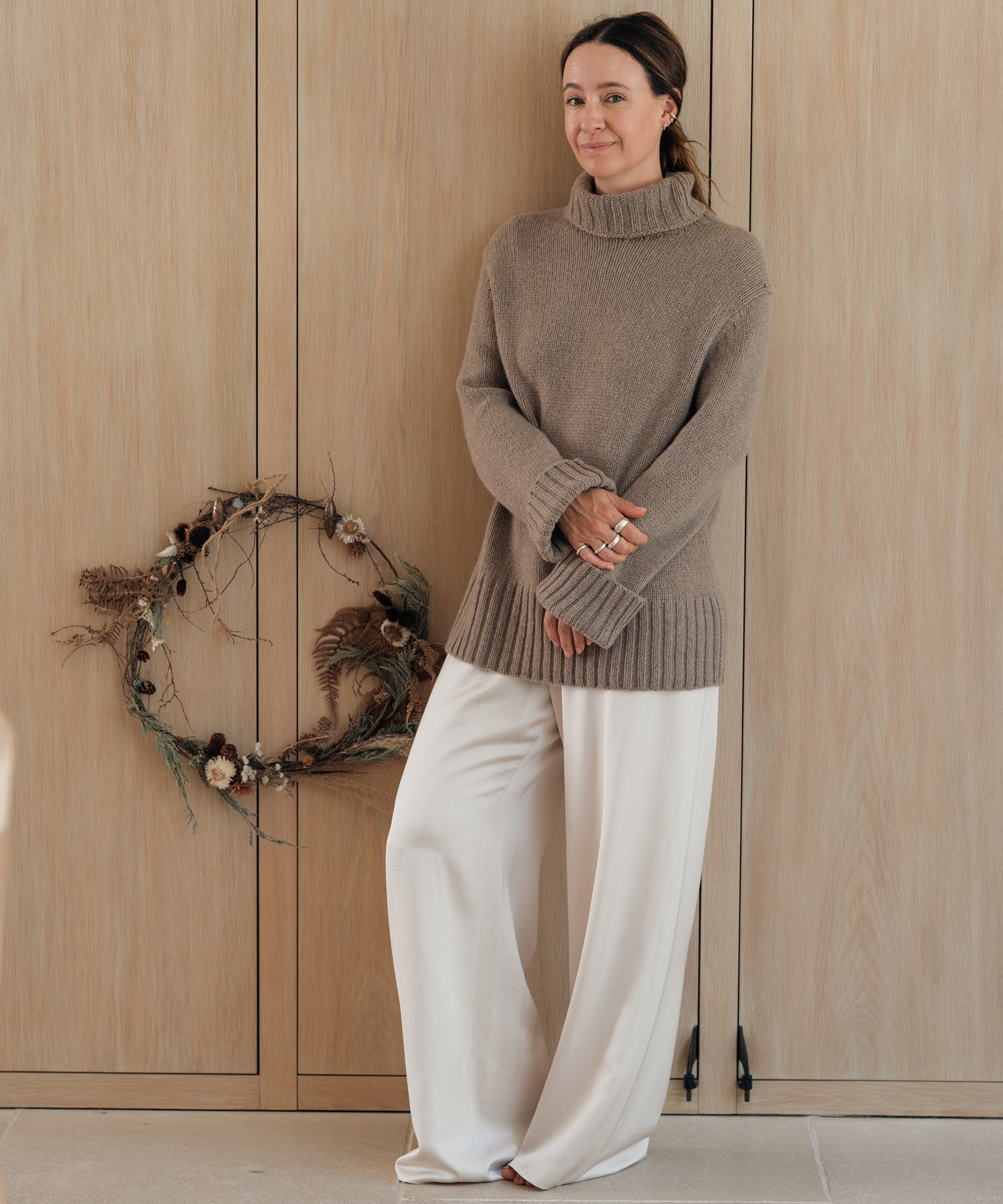jenni wearing a taupe cashmere turtleneck and silk pants standing next to a wreath