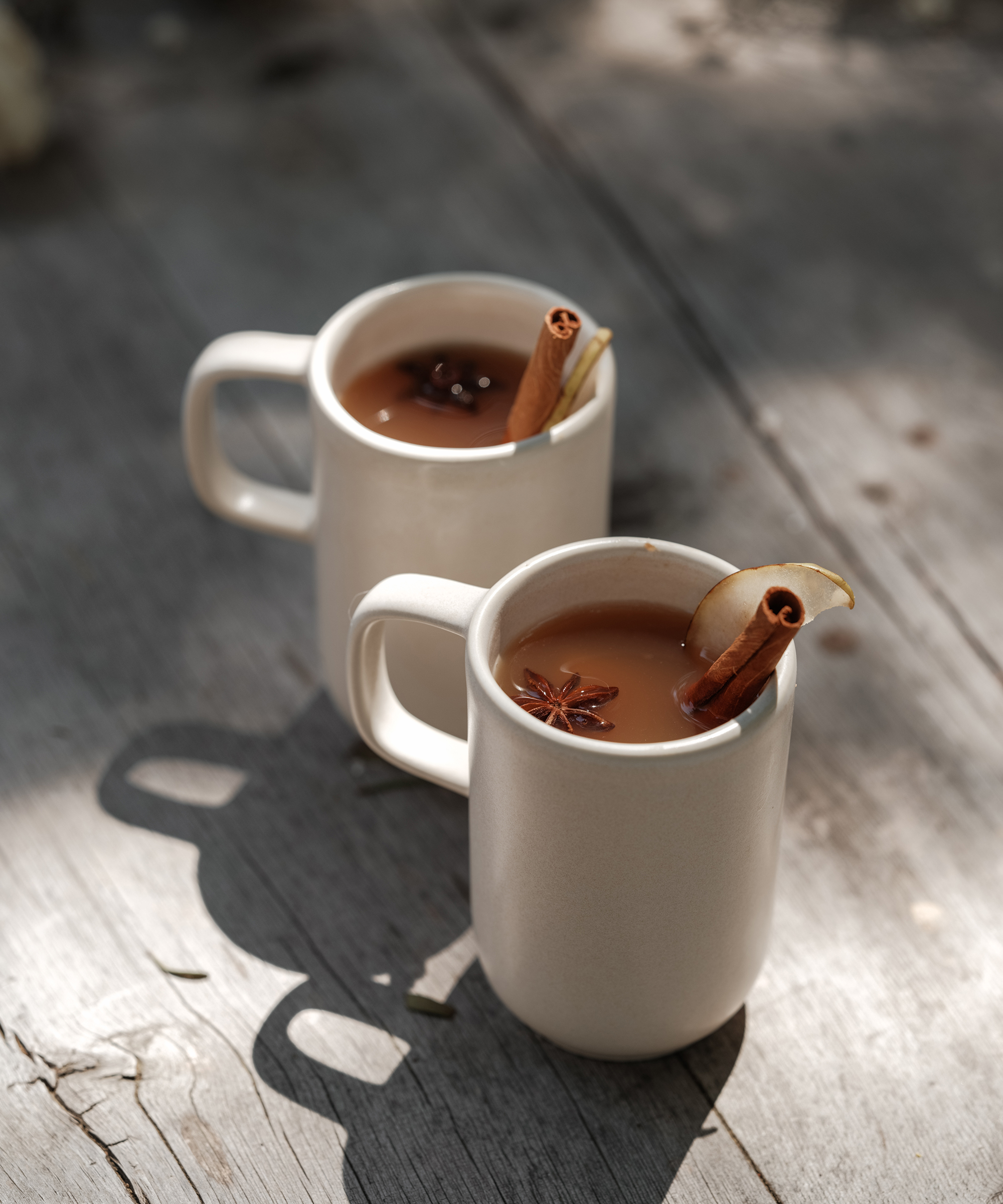 Two white mugs of spiced tea or cider sit on a wooden surface, each garnished with a cinnamon stick and star anise; one also has a slice of apple. Sunlight casts soft shadows beside these cozy companions to holiday desserts.