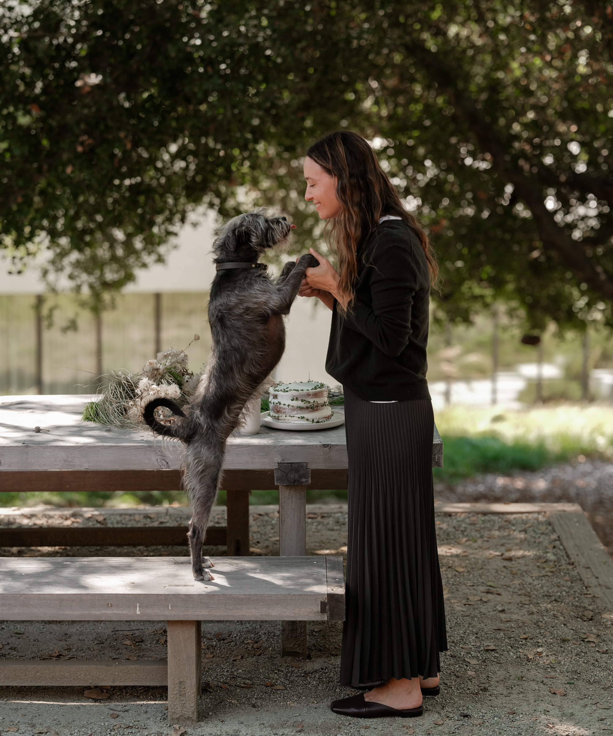 A woman stands by a picnic table, smiling and holding paws with a gray dog standing on its hind legs. Holiday desserts and flowers decorate the table, while trees provide shade in the background.