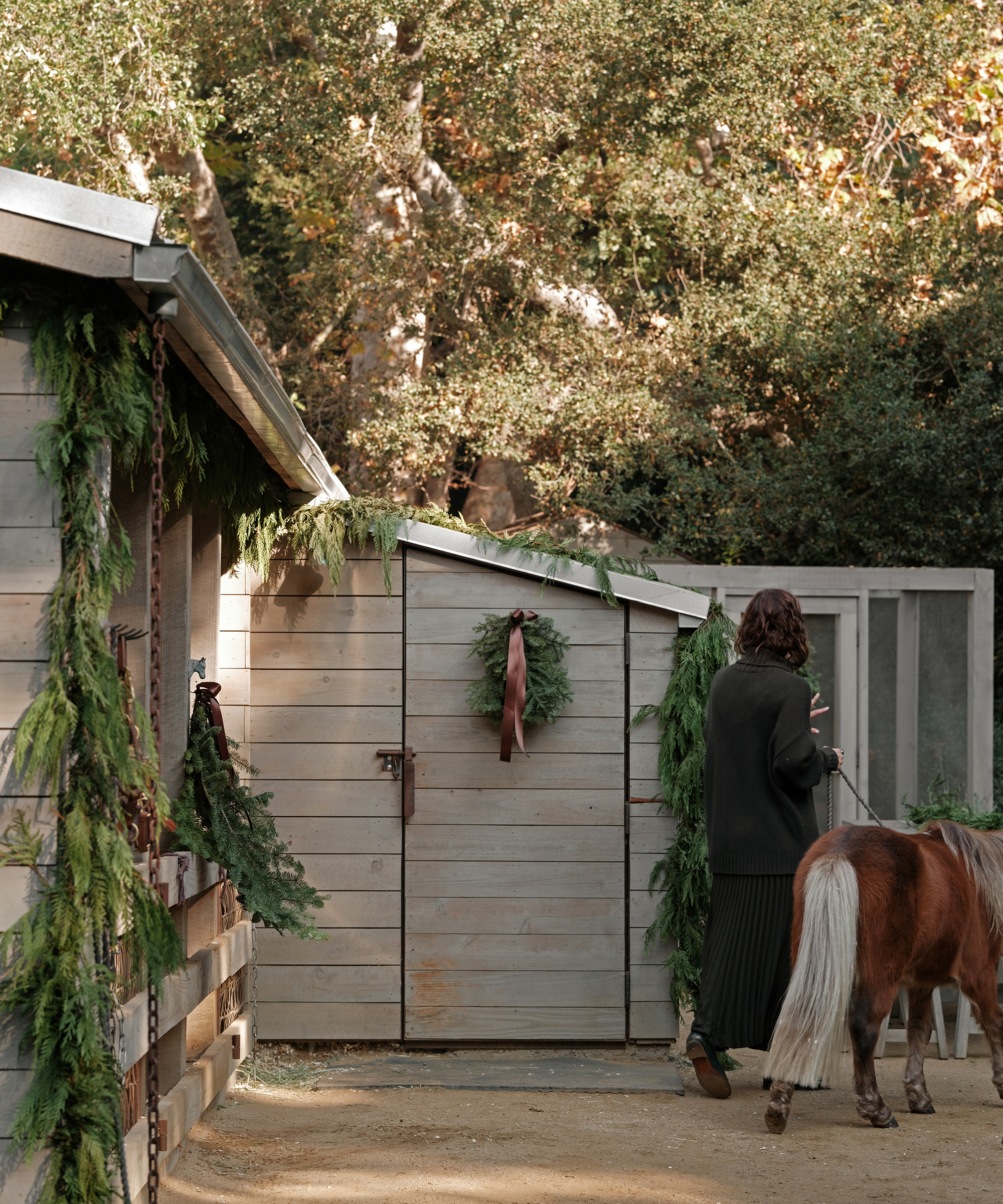 jenni wearing a pleated skirt and forest green cashmere sweater walking her horse by a stable