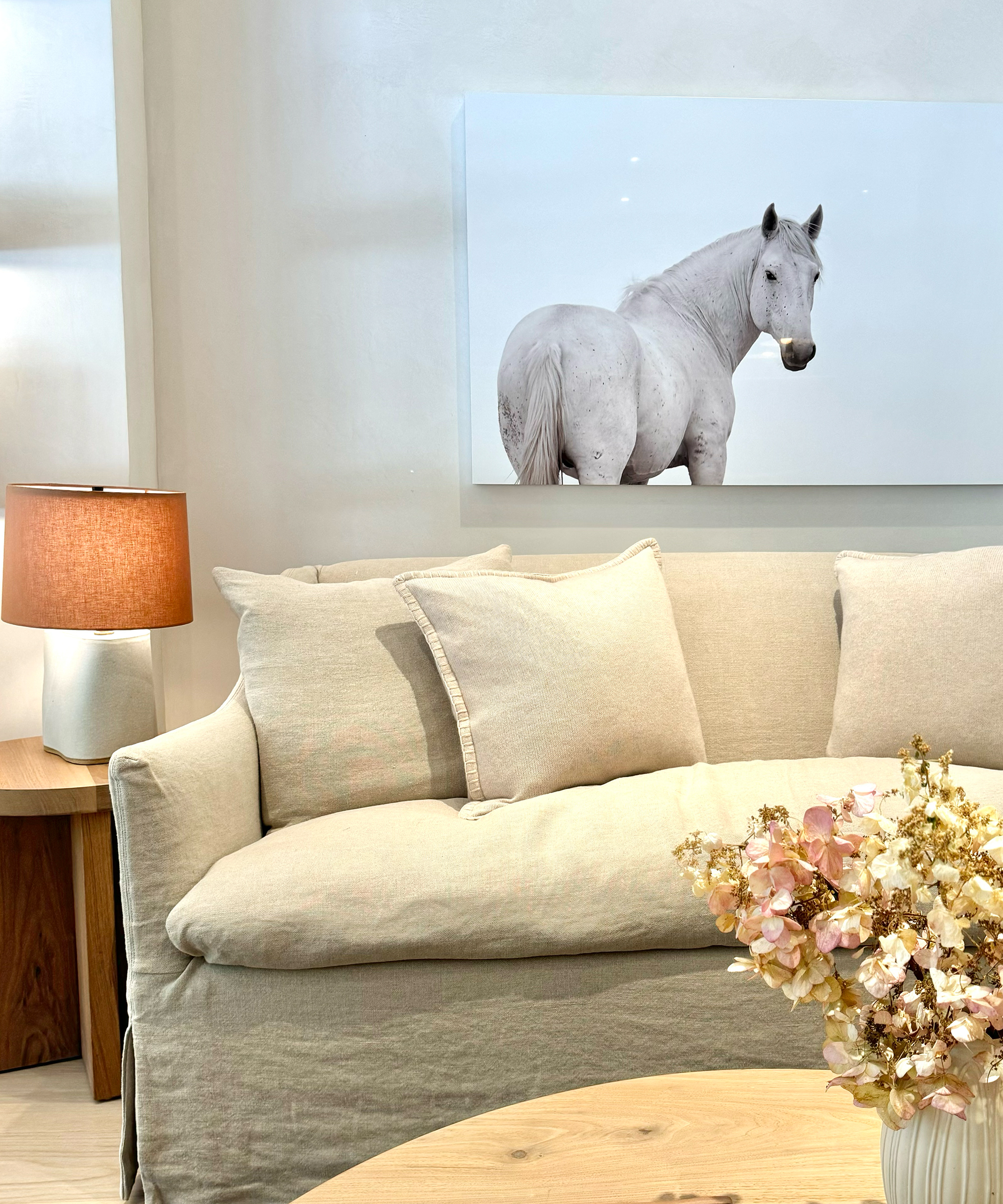 A beige sofa with cushions sits against a white wall, above which hangs a large photo of a white horse. On Giving Tuesday, the cozy setting is complete with a side table, lamp, and round wooden coffee table adorned with dried flowers.