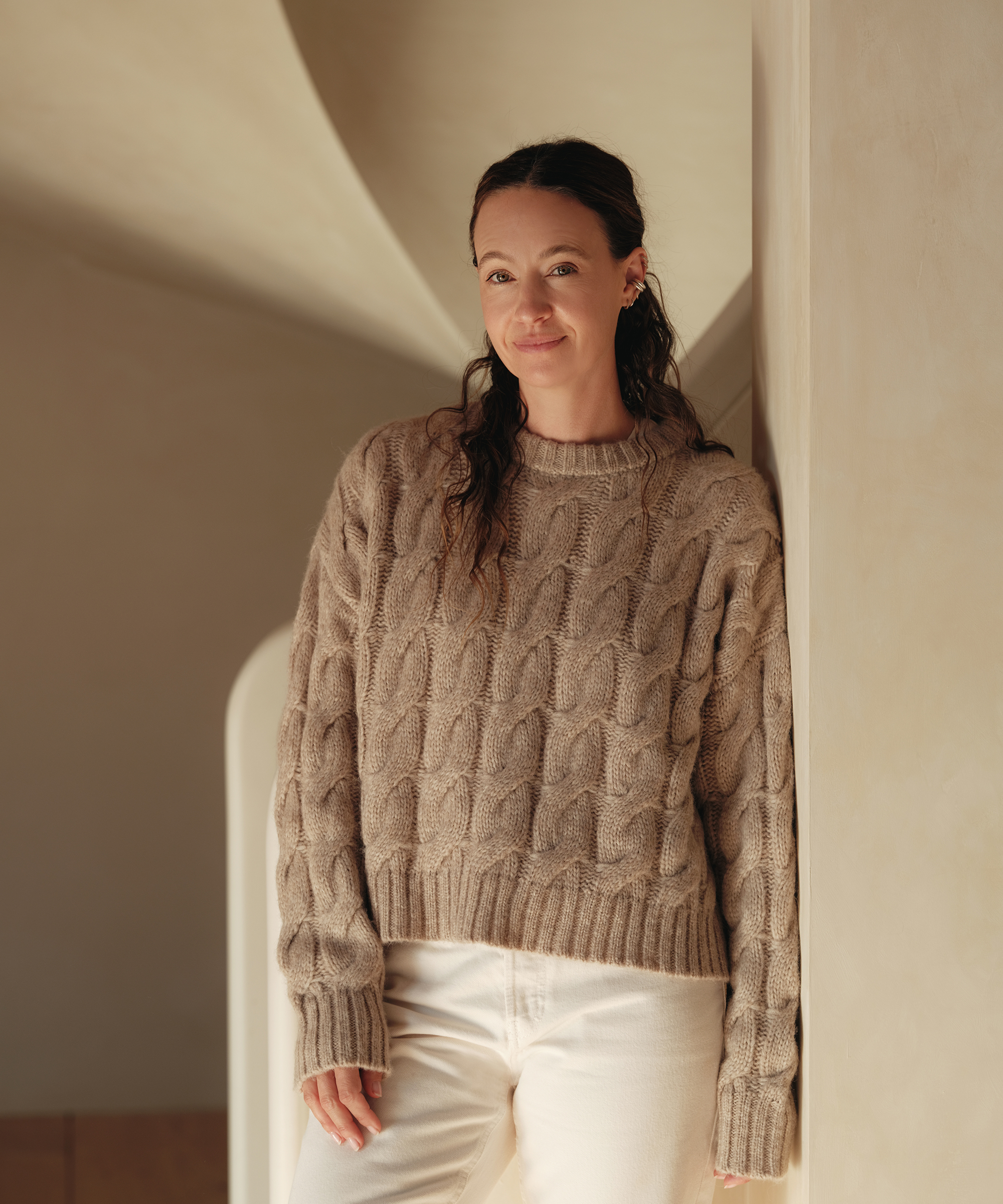 jenni wearing a thick cable knit sweater and creme jeans leaning against a wall