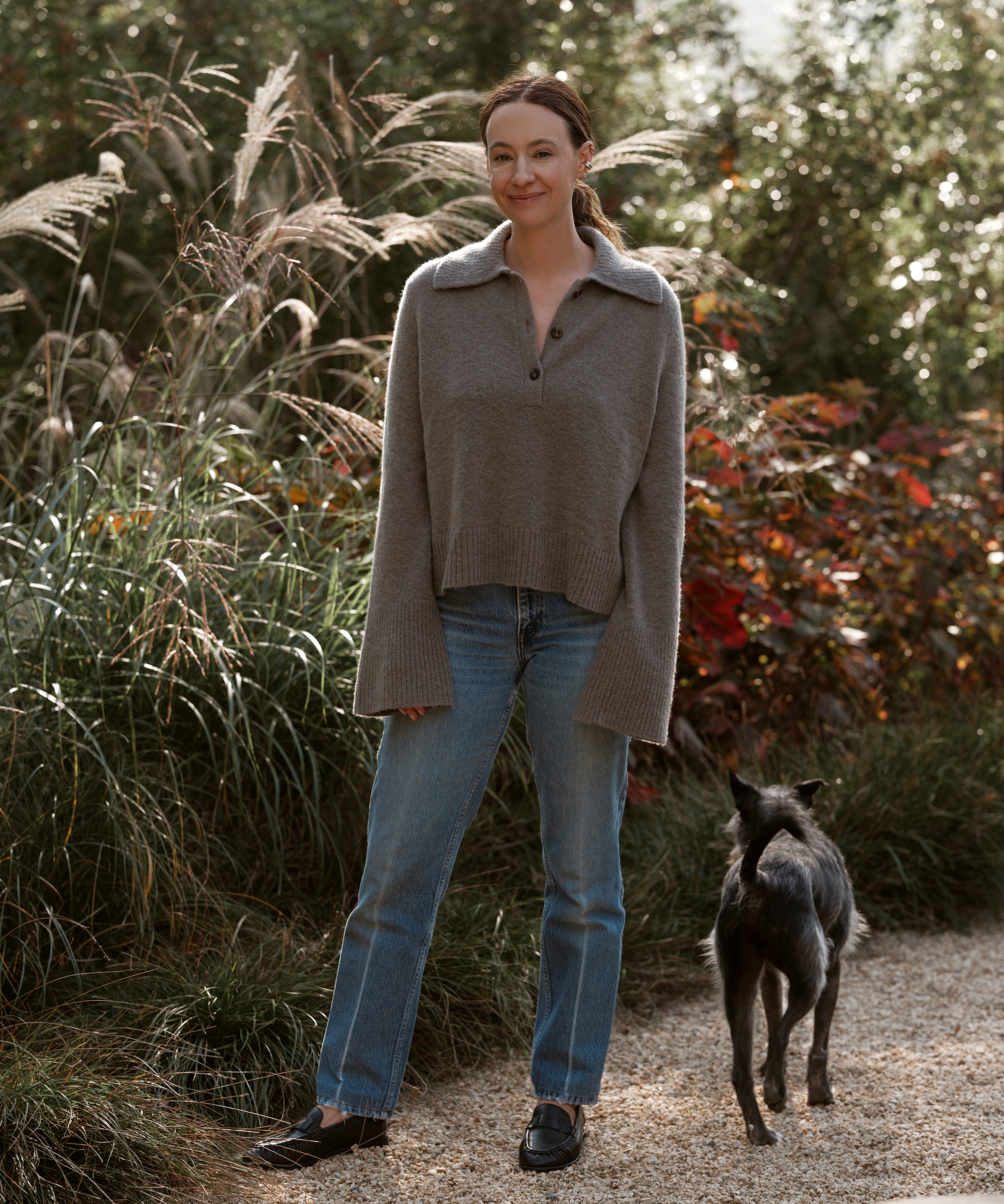jenni wearing a grey long sleeve button cashmere polo and jeans standing next to her dog in a gravel path