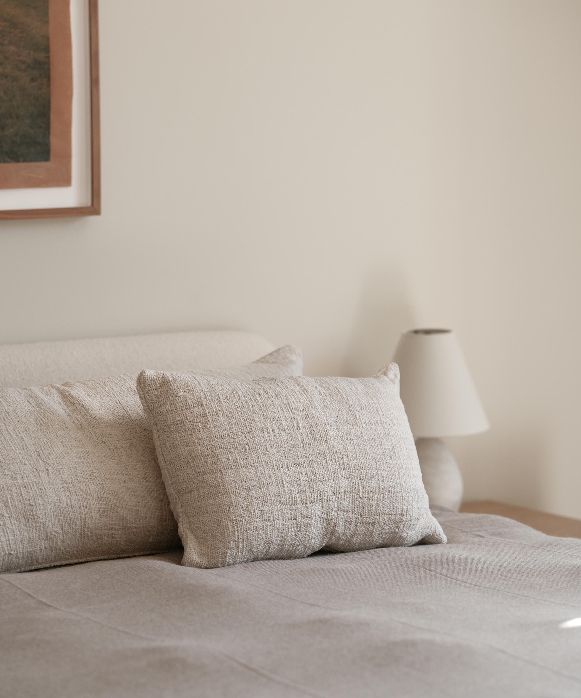 A neatly made bed with textured beige pillows and a taupe blanket, beside a small wooden nightstand with a white lamp—creating a cozy retreat worthy of Whitney Cummings. A framed artwork hangs on the light-colored wall above the bed.