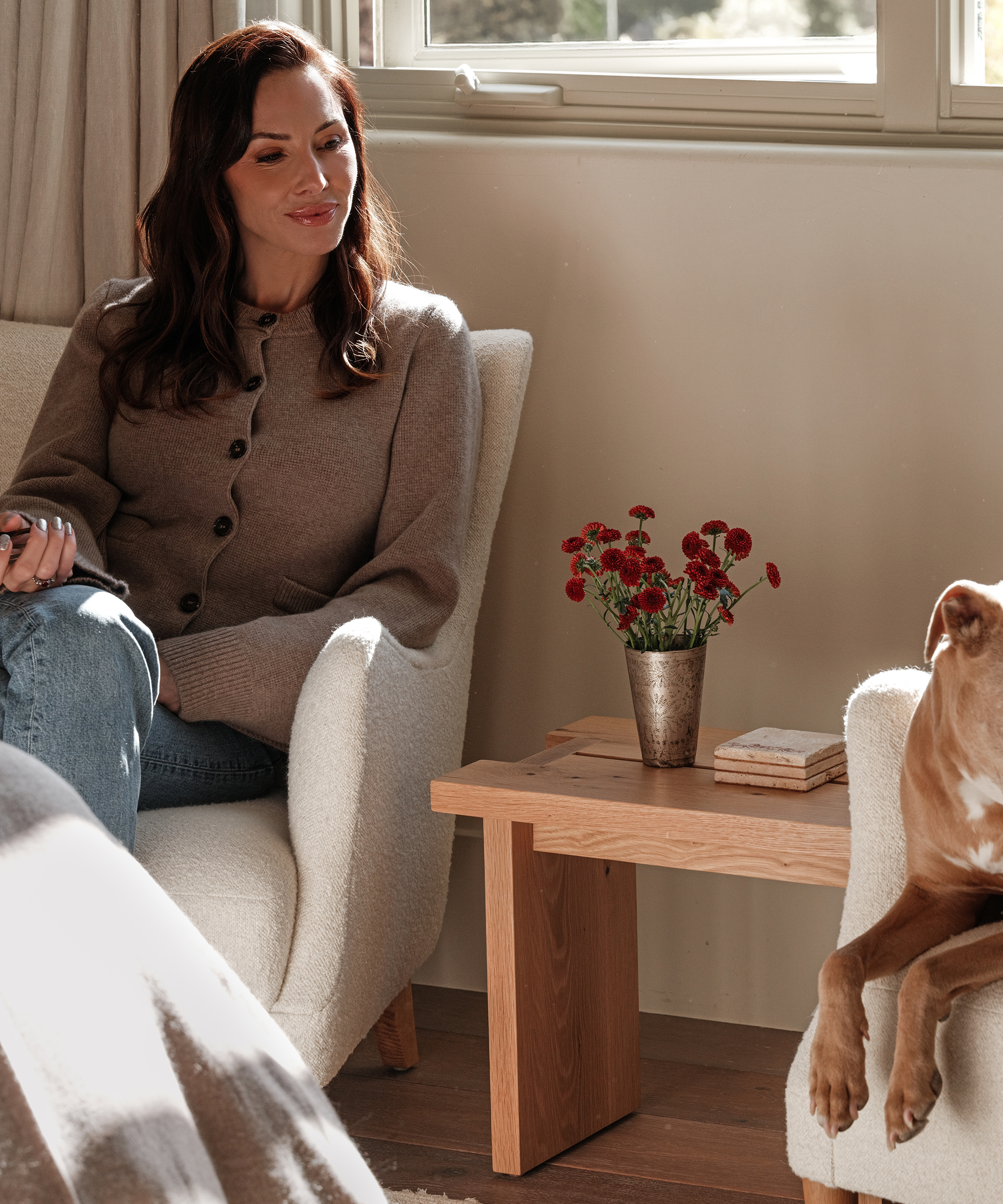 Whitney Cummings, in a brown sweater, sits on a cream armchair, smiling at a dog resting nearby. A small wooden table with a vase of red flowers and coasters sits between them as sunlight streams through the window.