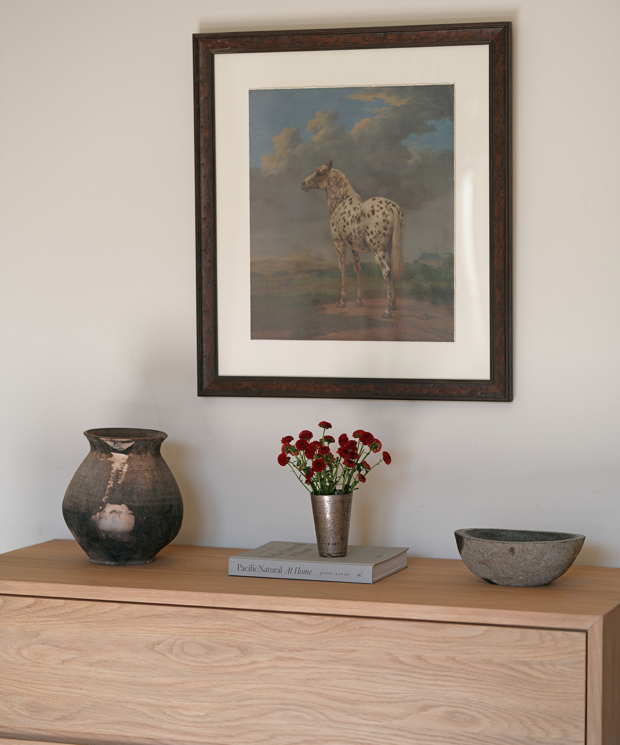 A wooden dresser displays a dark ceramic vase, a metal cup with red flowers, a book, and a stone bowl. Above it hangs a framed painting of a spotted horse in a landscape—a scene reminiscent of Whitney Cummings’ eclectic style.