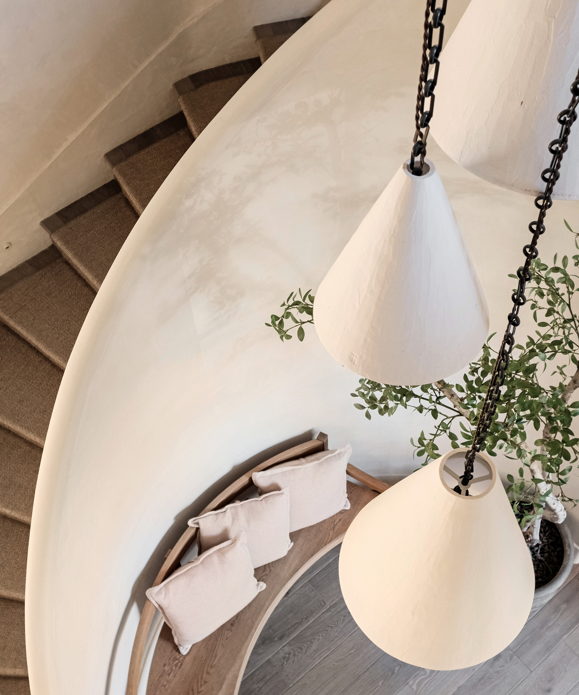 A top-down view of a curved staircase with beige carpet, a bench with three light-colored pillows beside it, large white hanging pendant lights, and a leafy green plant—exuding the soft, minimalist aesthetic Whitney Cummings loves.