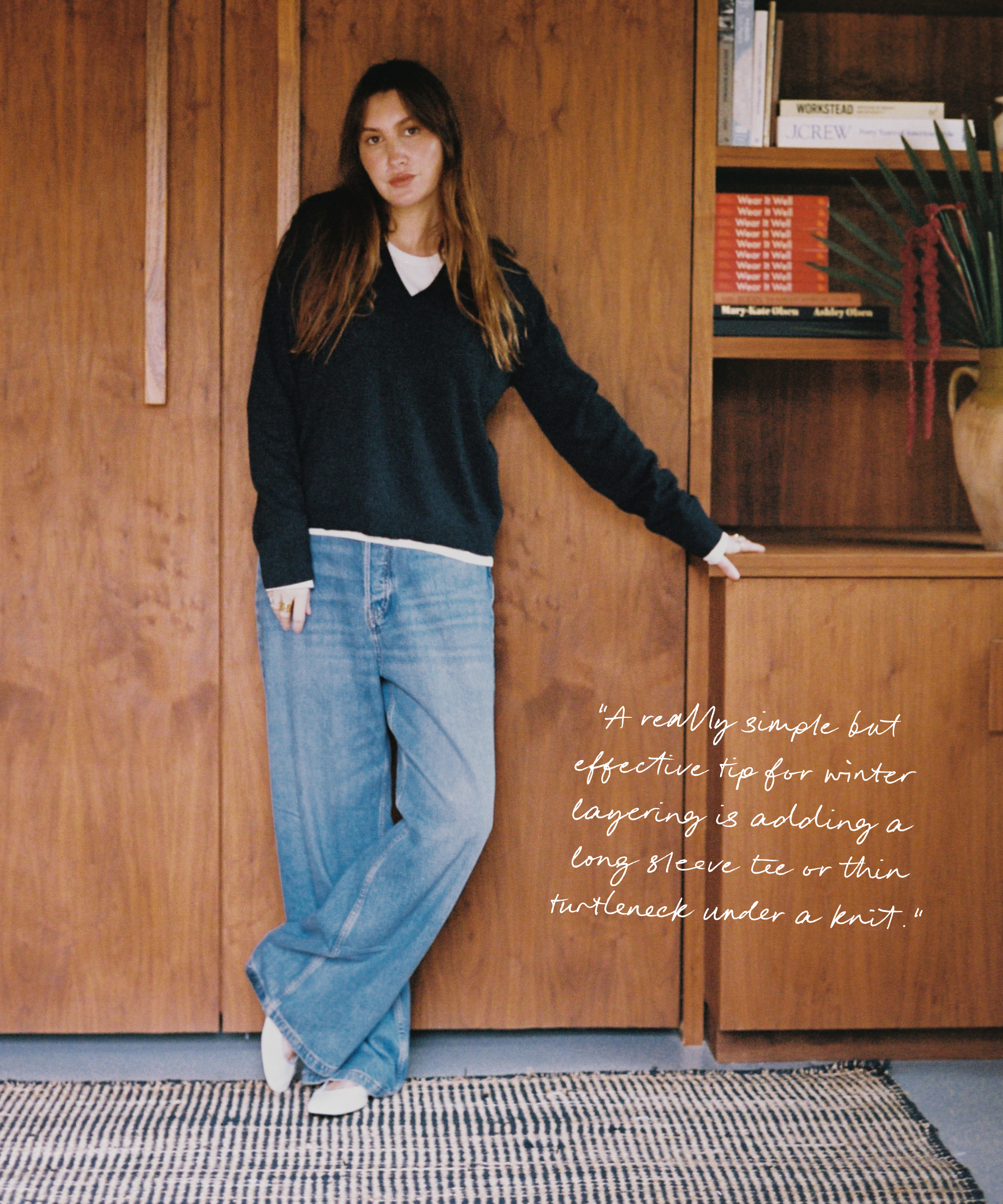 A woman stands indoors by a wooden cabinet, channeling Allison Bornstein’s style in a black sweater over a white shirt, wide-leg blue jeans, and white shoes. Text beside her shares winter layering tips. A bookshelf with books and plants is in the background.