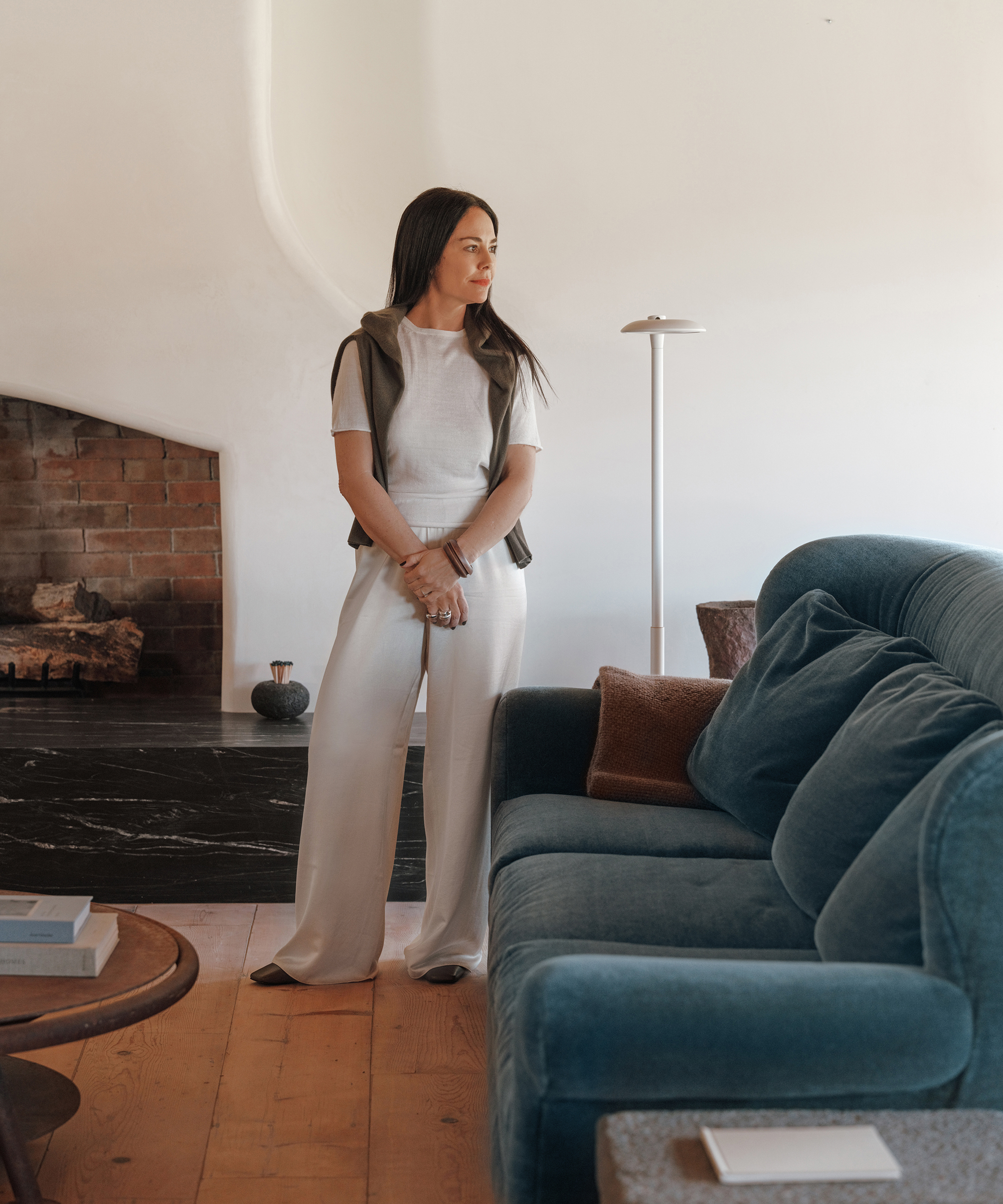 corinne standing in a living room next to a blue velvet couch wearing ivory silk pants and a white linen tee