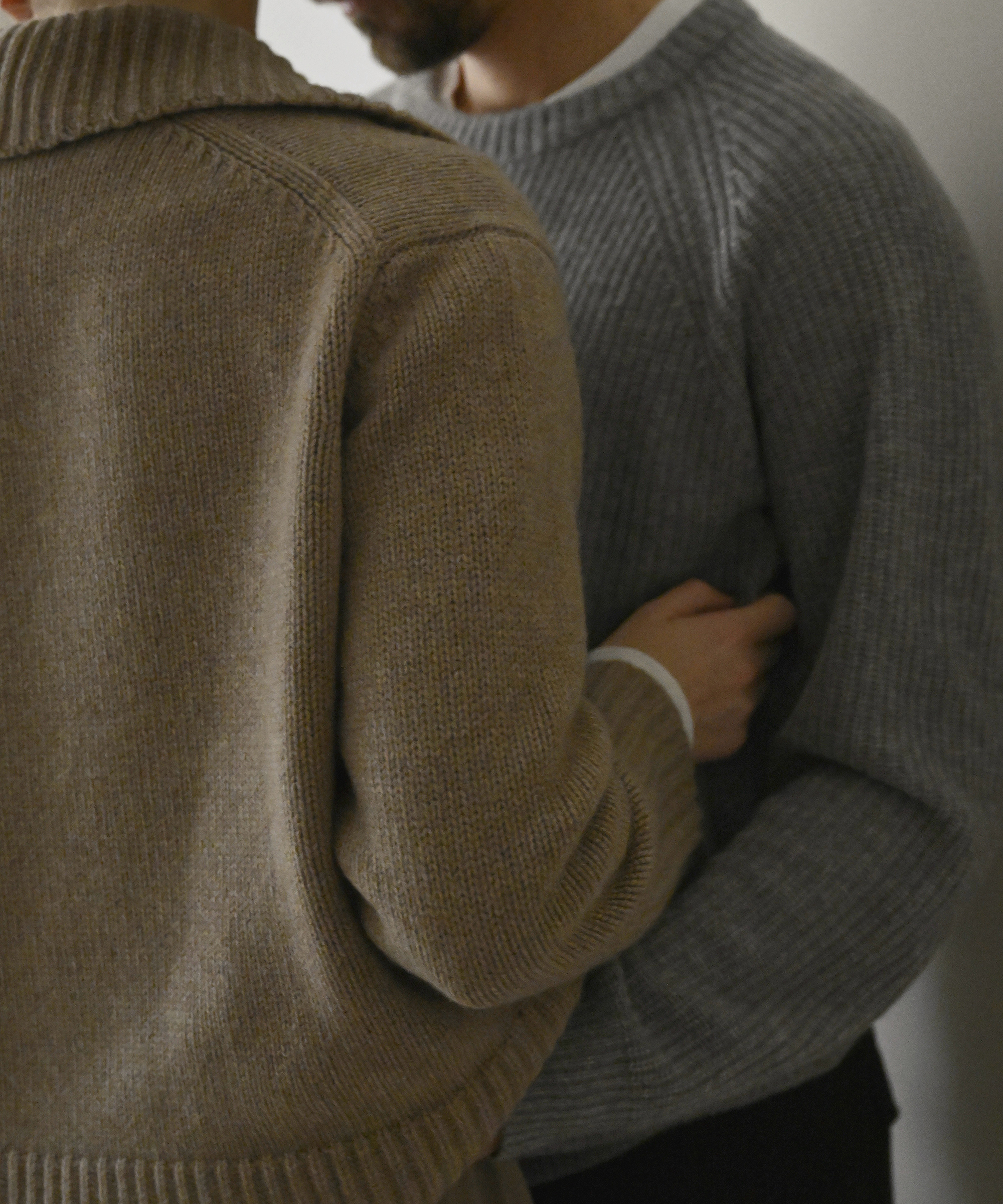 Two people stand close together indoors, both wearing knit sweaters—one in beige and the other in gray. Channeling Ellen Claesson’s signature cozy style, the person in beige gently holds the others side, creating an intimate and comforting atmosphere.