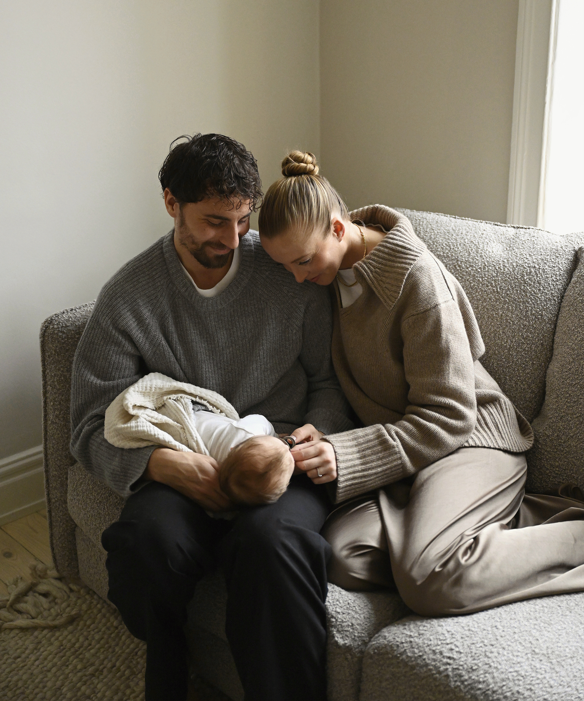 a couple wearing jenni kayne sweaters sitting on a couch holding their baby
