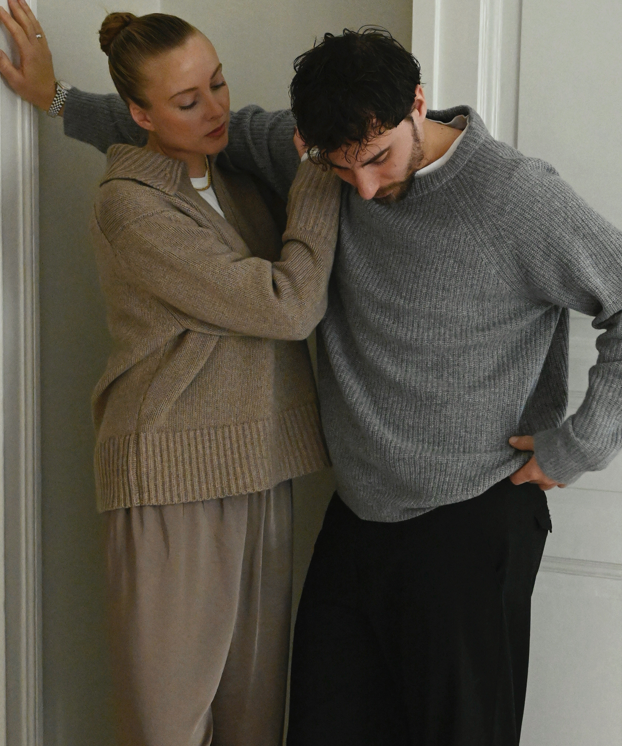 a couple leaning against a wall wearing an oatmeal sweater and silk pants and a man in a grey cashmere sweater