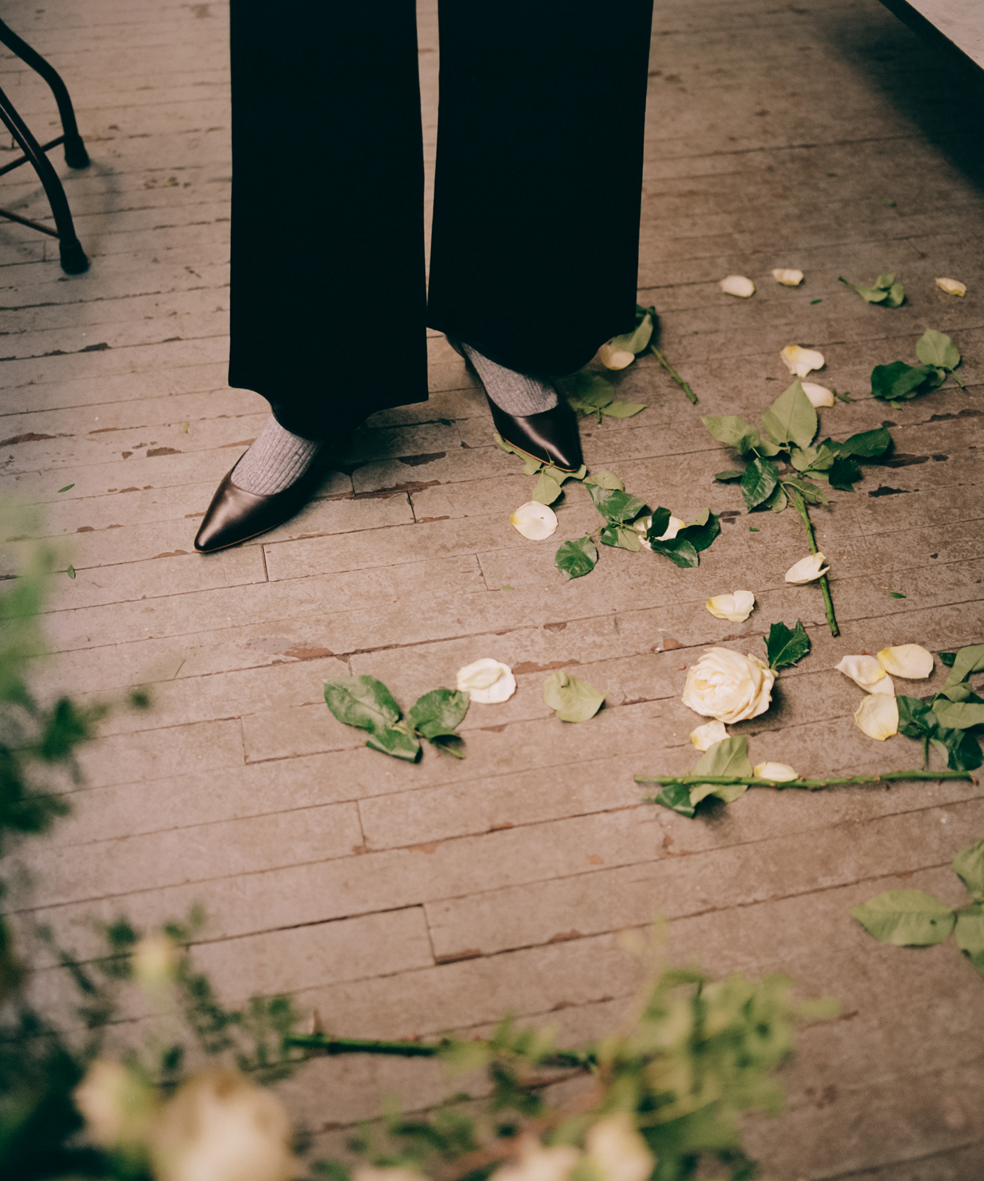 A person wearing dark pants and black heeled shoes stands on a wooden floor scattered with yellow roses and green leaves—an elegant scene reminiscent of Flowers by Ford.