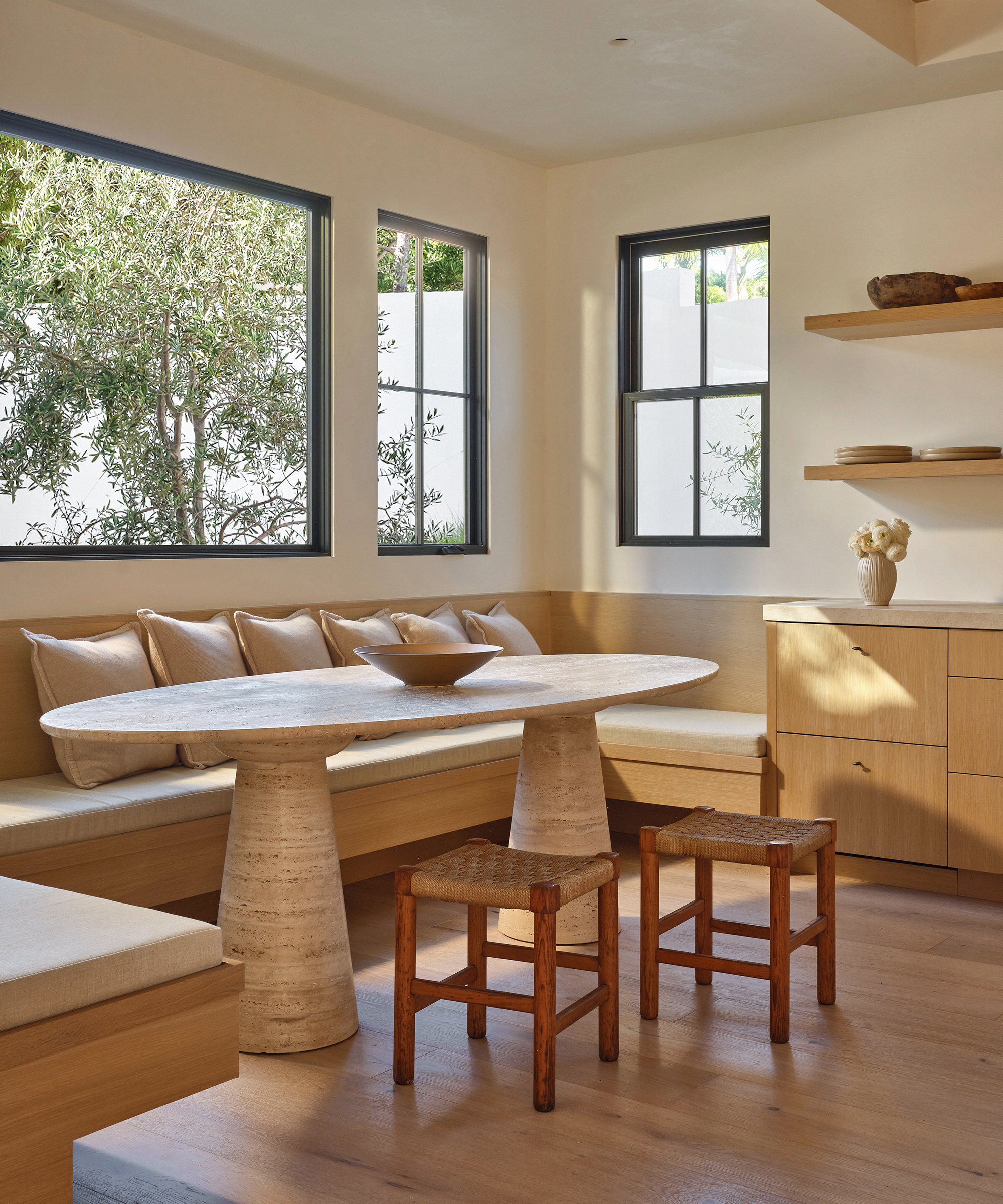 A bright, modern dining nook in Manhattan Beach with a round stone table, two wooden stools, built-in bench seating with tan cushions, wood floors, and large windows letting in natural light. Minimalist shelves hold a vase and plates.