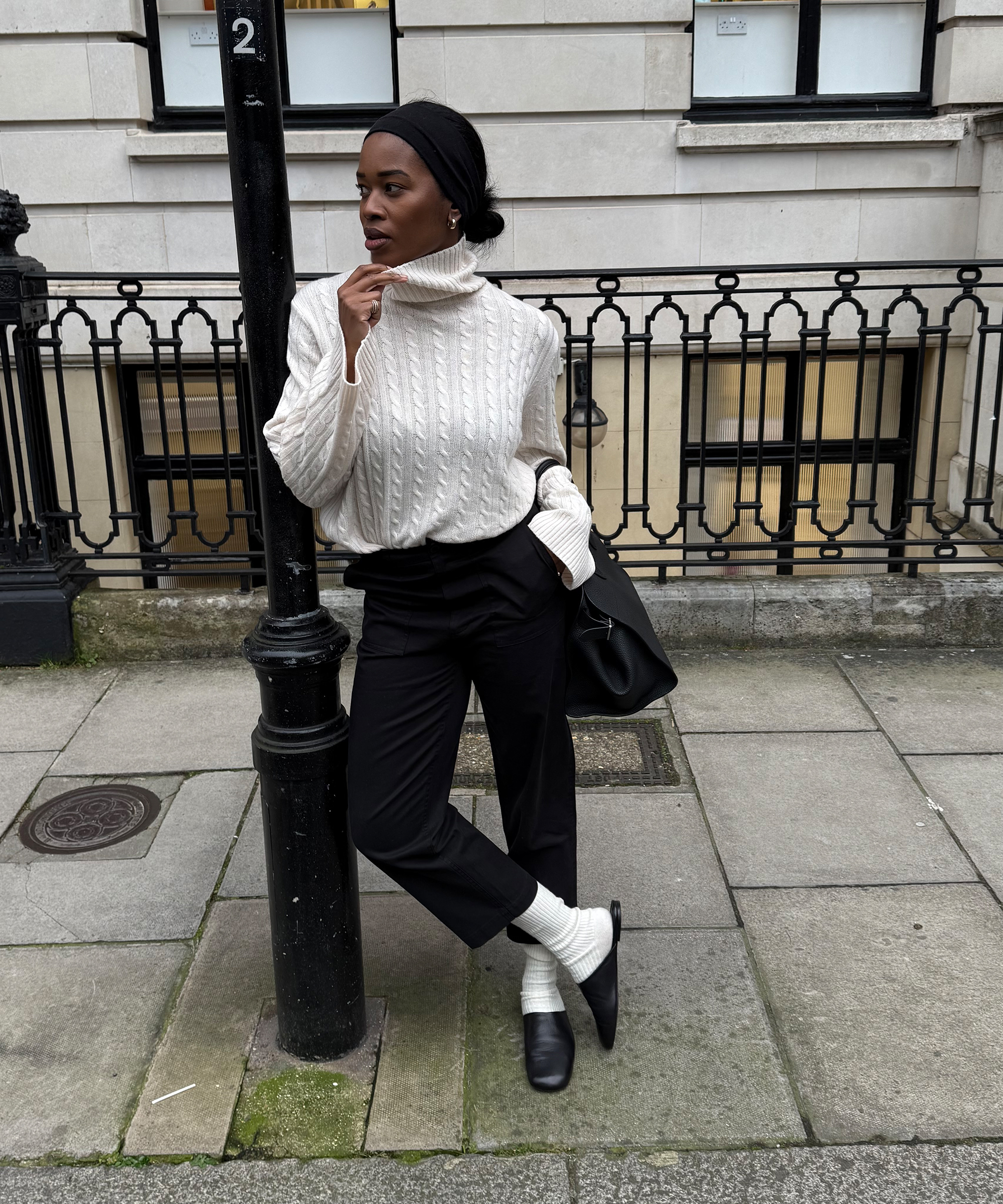 A woman in a cream turtleneck sweater, black pants, white socks, and black loafers leans against a streetlamp on a city sidewalk, looking to the side with one hand touching her chin.