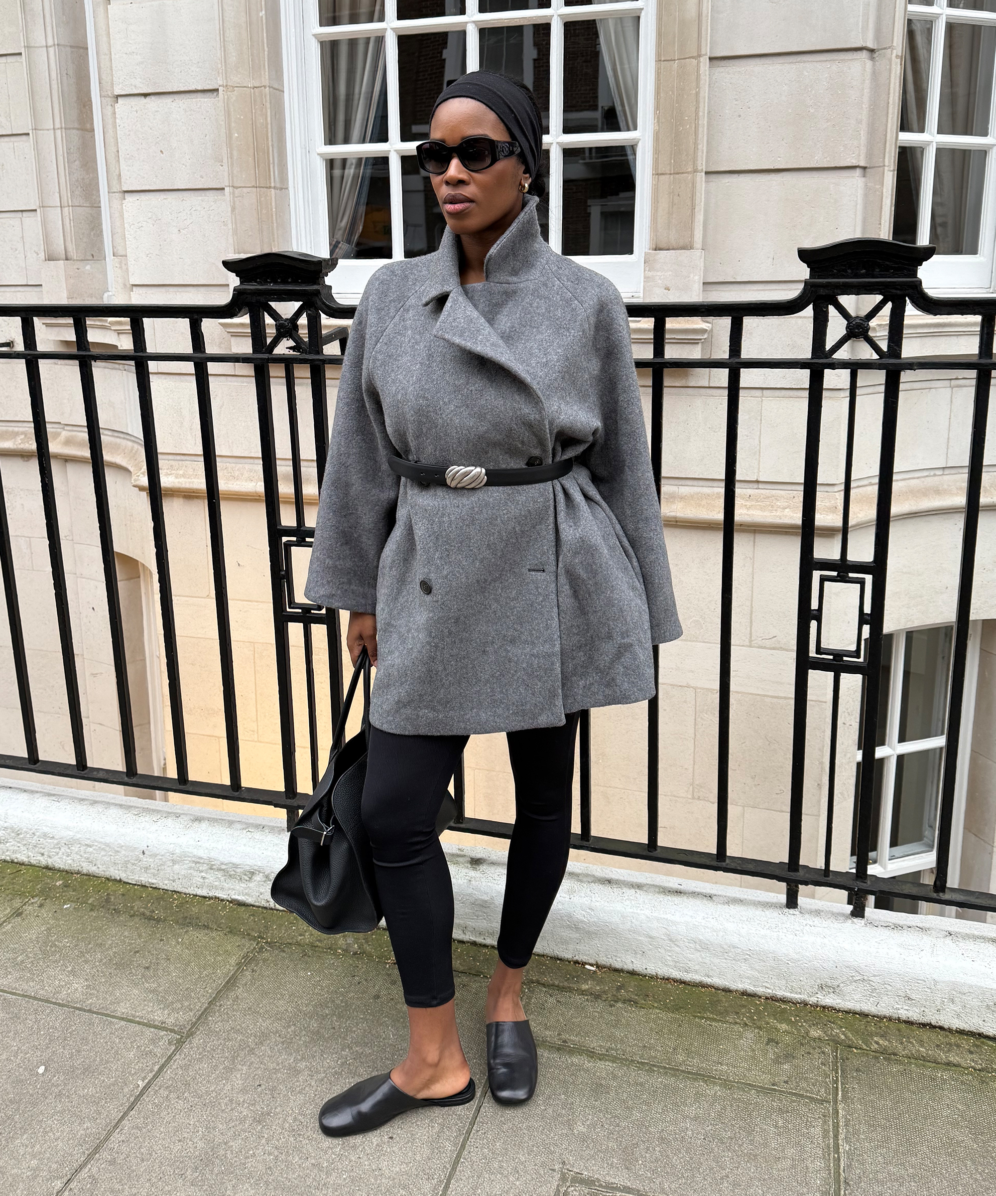 A woman stands on a sidewalk in front of a black railing, wearing a grey coat cinched with a belt, black leggings, black slip-on shoes, a headscarf, sunglasses, and holding a black handbag.