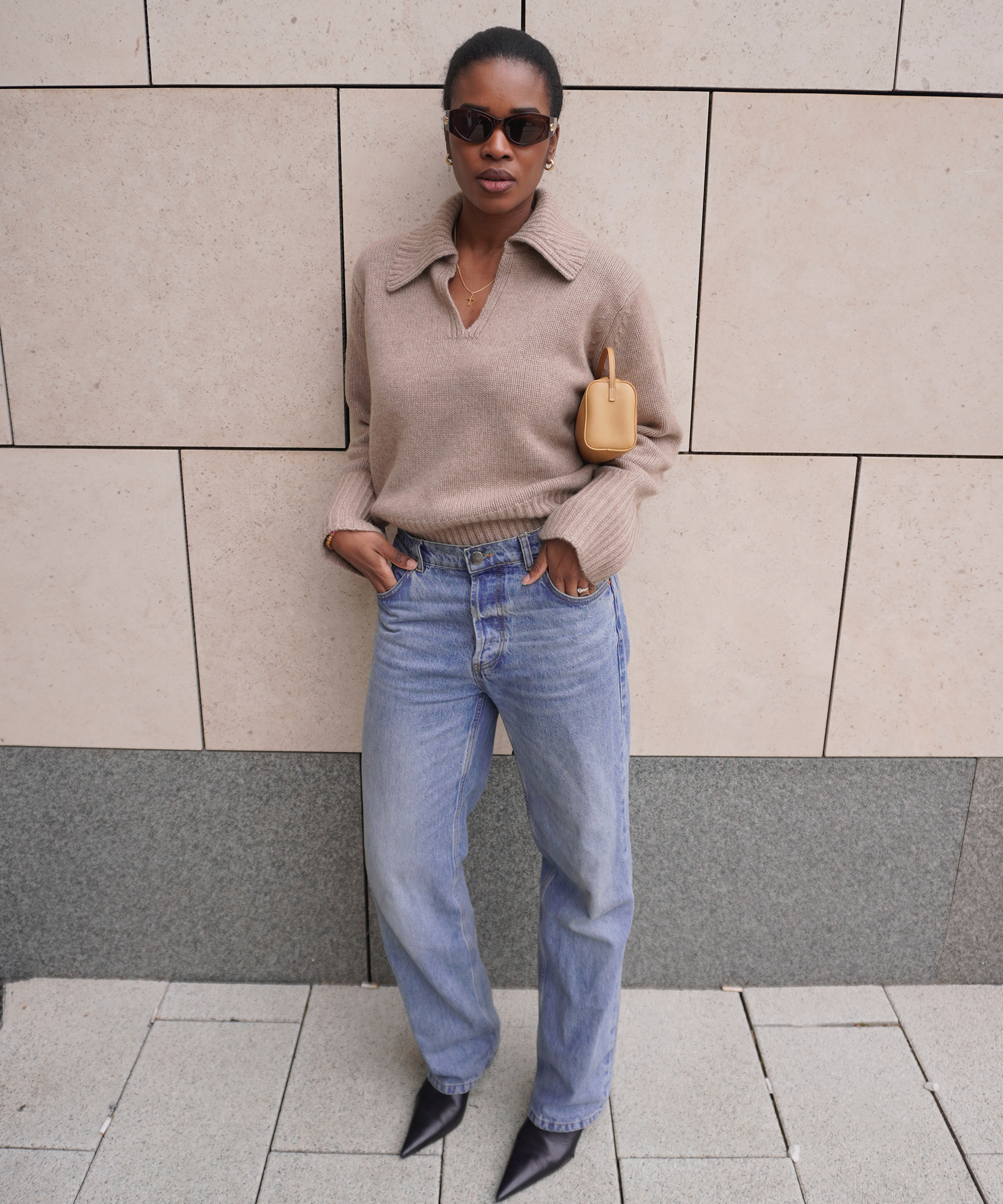 A woman wearing sunglasses, a beige sweater with a collar, light blue high-waisted jeans, and black pointed shoes stands against a tiled wall. She has a small tan bag tucked under her arm.