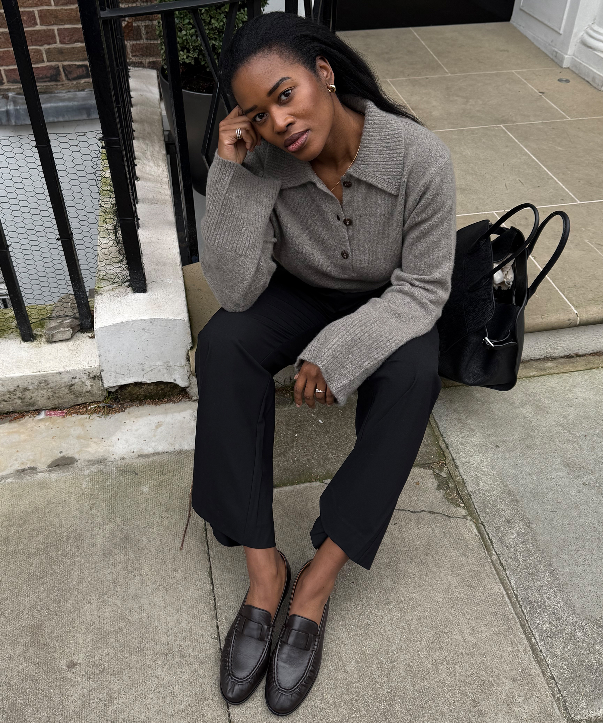 A woman sits on a stone step, resting her elbow on her knee and her head on her hand. She wears a taupe sweater, black pants, and brown loafers. A black bag sits beside her. She looks thoughtfully at the camera.