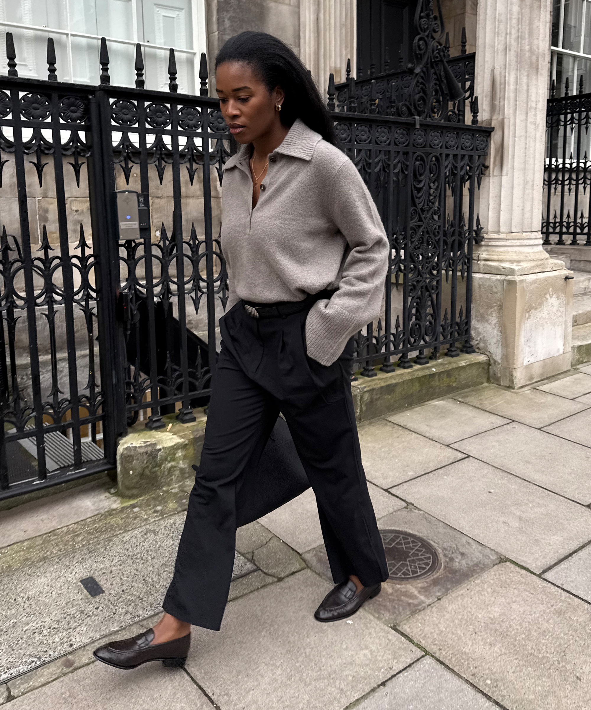 A woman walks on a city sidewalk in front of ornate black iron railings, wearing a gray collared sweater, black pleated trousers, and dark loafers. She has long hair and her hands are in her pockets.