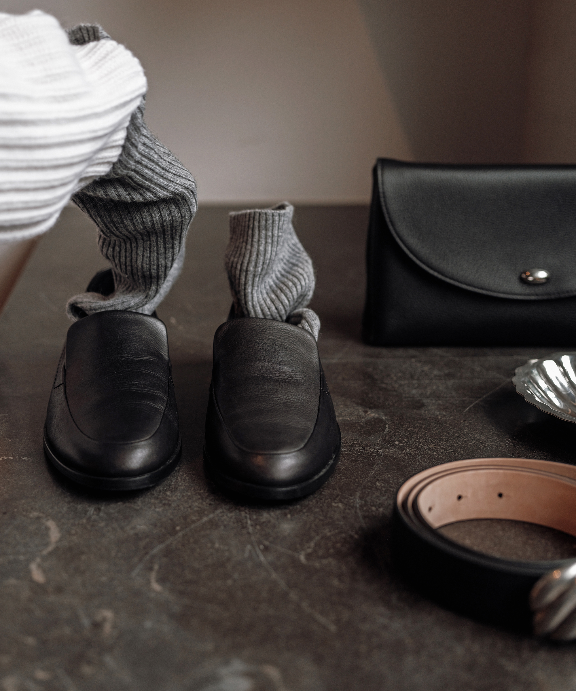 A pair of black leather loafers with gray socks inside, a black leather wallet, and a brown leather belt arranged on a dark surface exemplify The Art of Layering According to Julia Rabinowitsch.