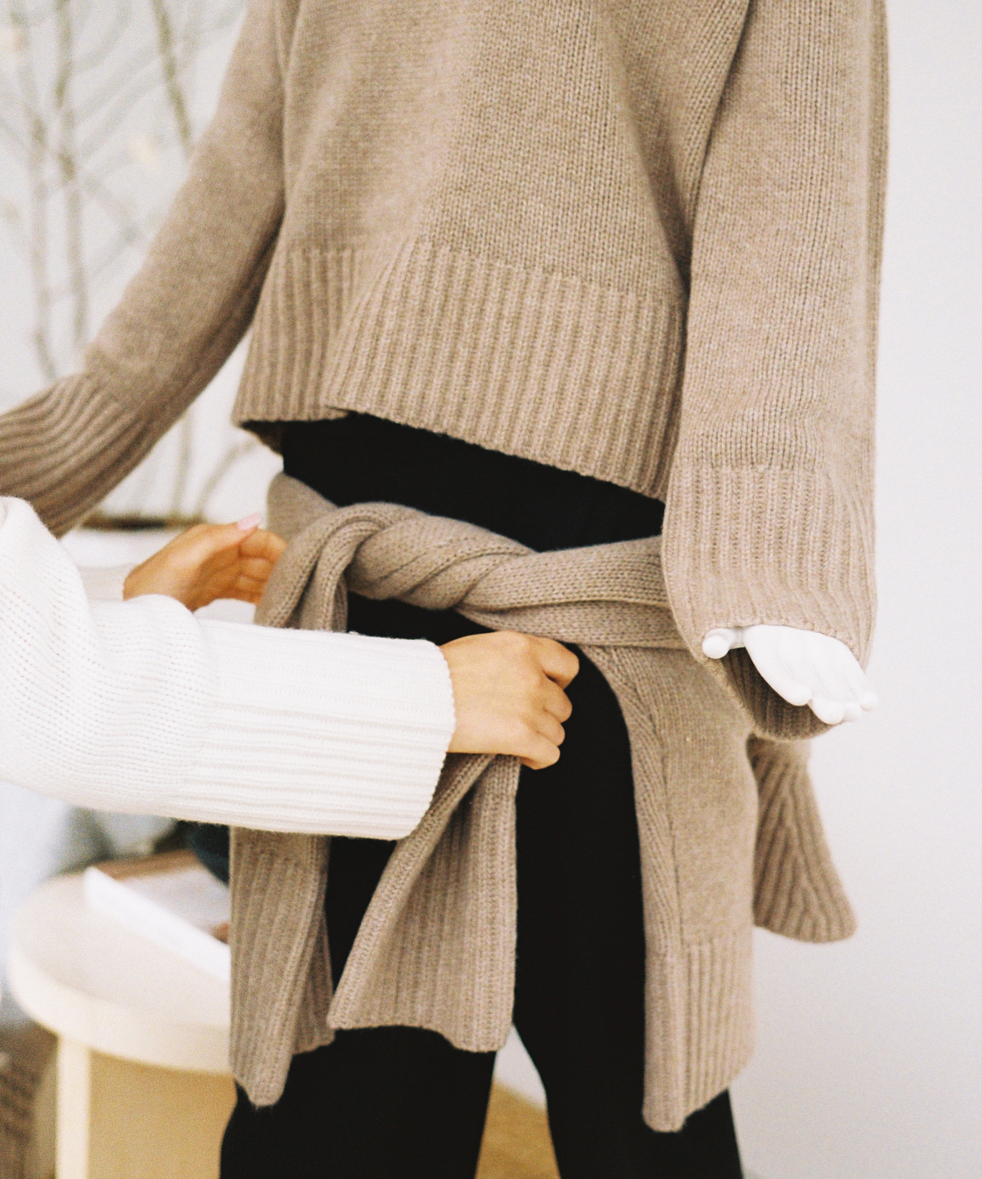 In The Art of Layering According to Julia Rabinowitsch, a person ties a beige knit sweater around another, both in matching sweaters and black pants with long sleeves—an effortless indoor styling moment.