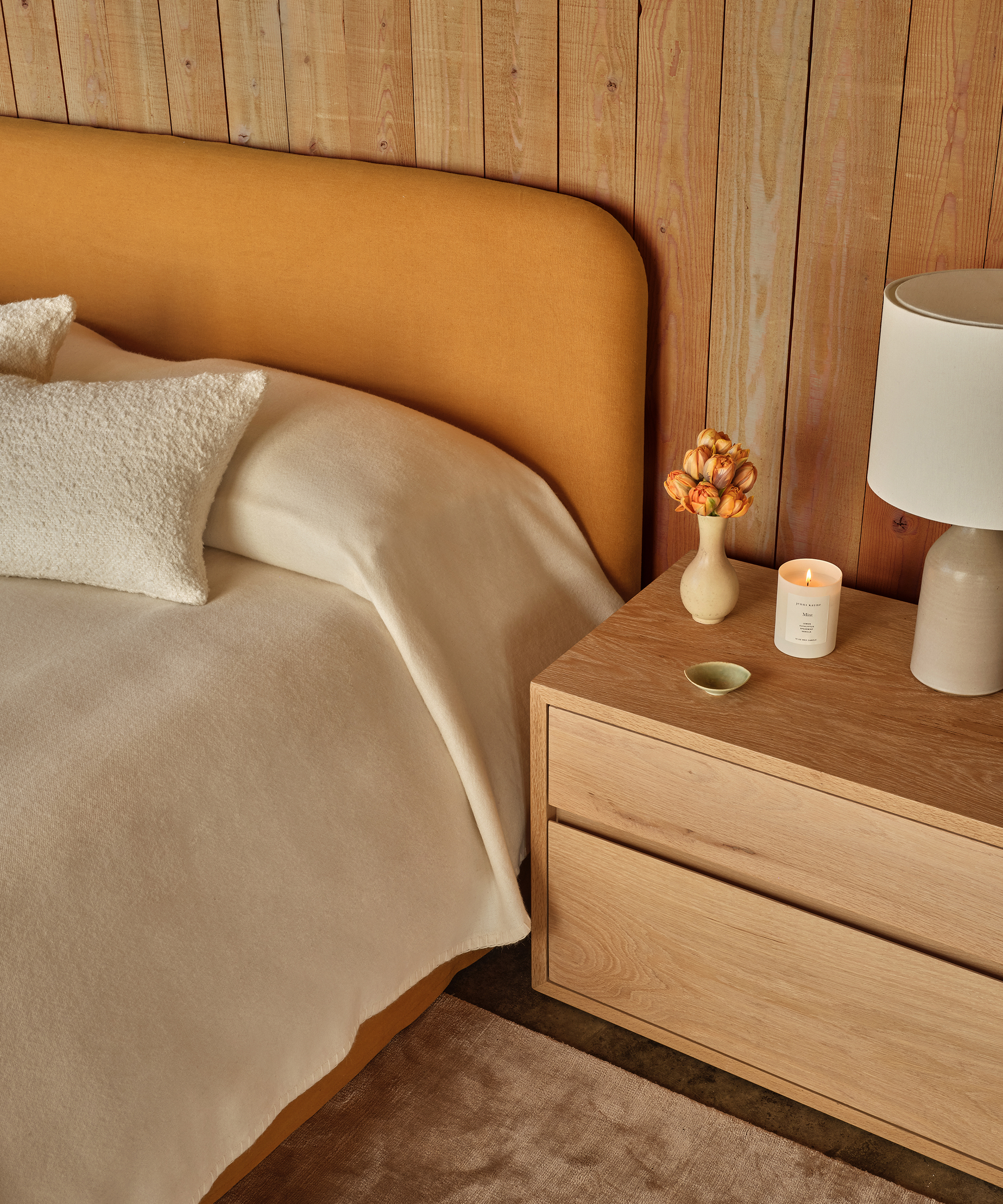 A cozy bedroom with a mustard yellow upholstered bed, cream bedding, and a white textured pillow captures the charm of spring decorating. The light wood nightstand holds a lamp, vase of flowers, candle, and a small decorative dish.