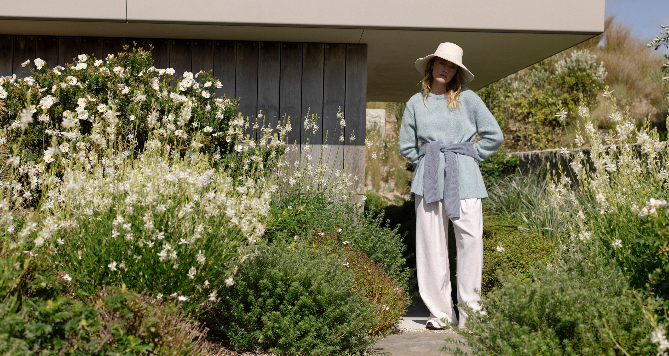 A woman wearing a wide-brimmed hat, light blue sweater, and white pants stands on a garden path surrounded by lush green plants and white flowers in front of a modern house, offering a behind the scenes glimpse of serene outdoor style.