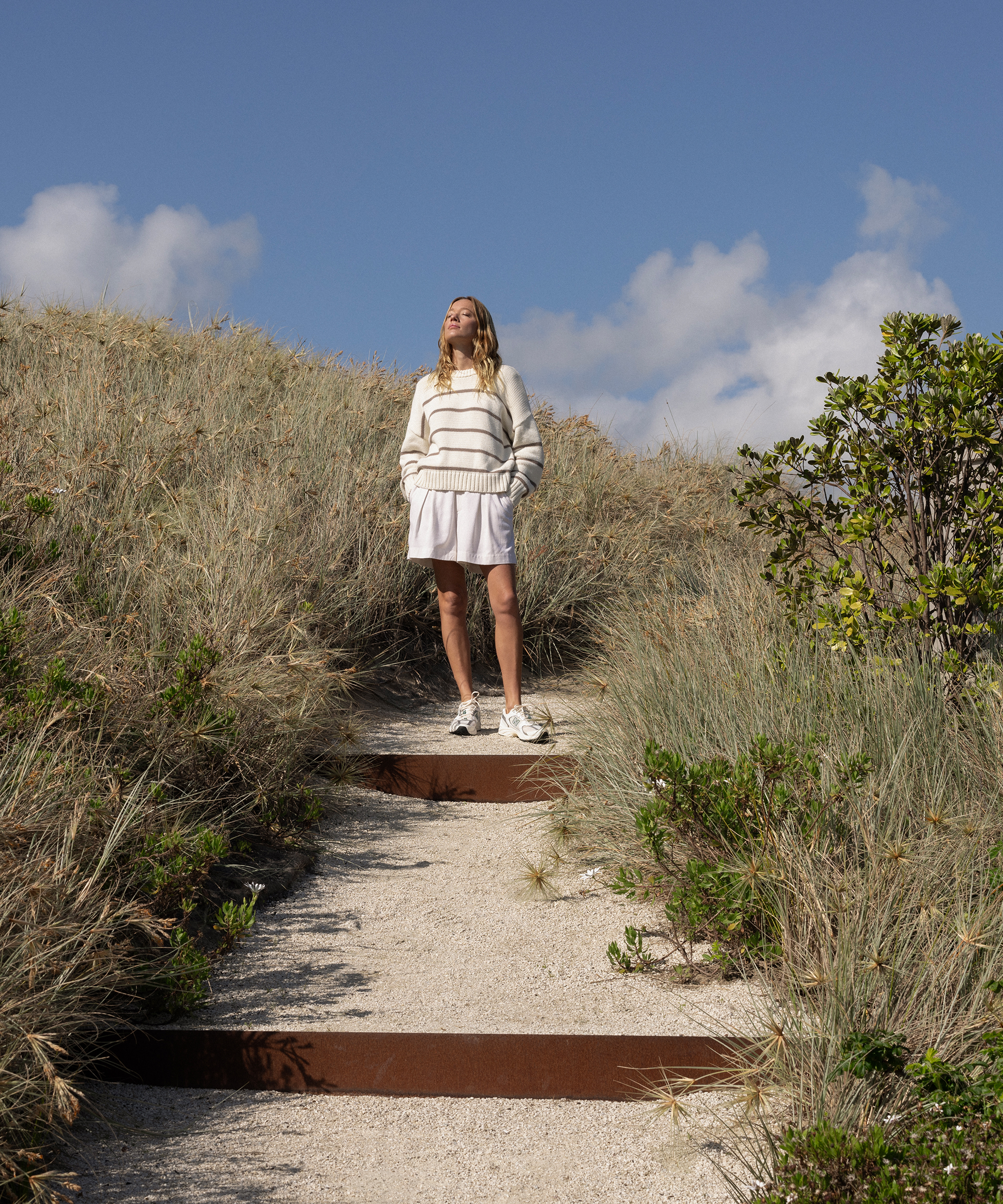 Behind the scenes, a woman in a white sweater and skirt stands on sandy steps, surrounded by tall grass under a blue sky with wispy clouds. She gazes upward, hands tucked in her pockets.