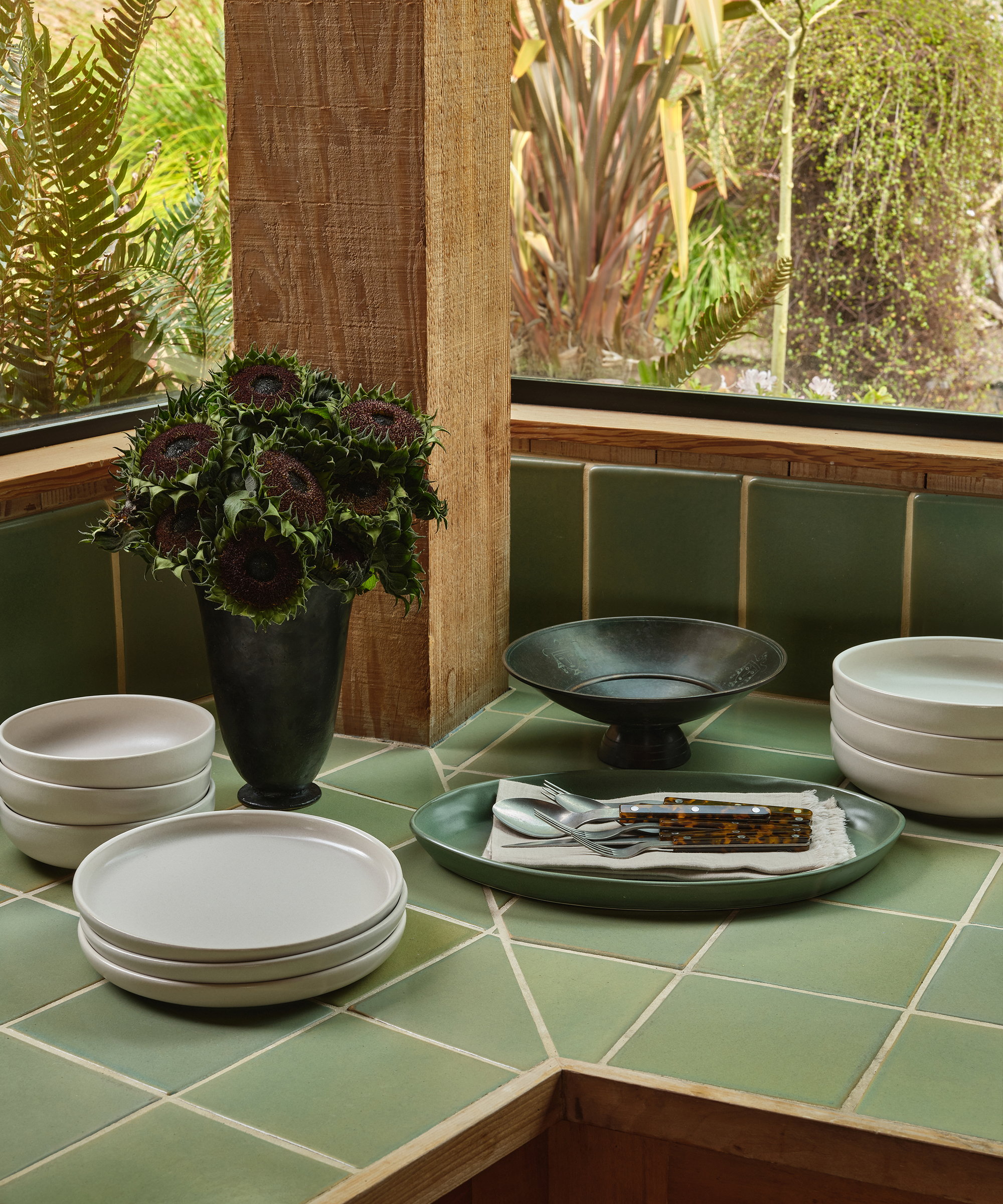 A kitchen corner perfect for spring decorating, featuring green tiled countertops, stacked white plates, a dark vase with flowers, a black pedestal bowl, and a green tray with cutlery by large windows overlooking a lush garden.