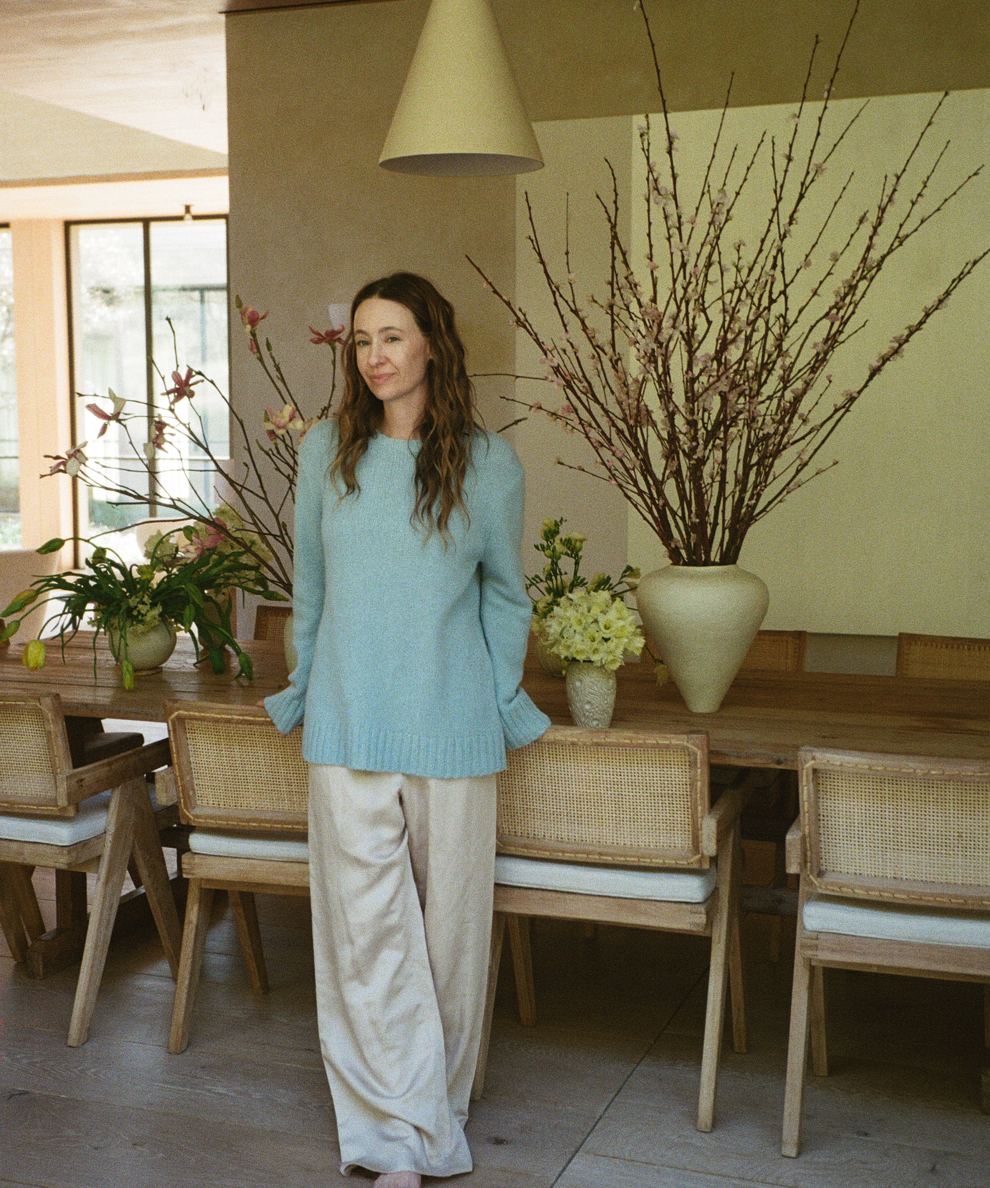 A woman in a light blue sweater and beige pants stands behind the scenes in a dining room with a wooden table, surrounded by large vases of flowers and branches, as natural light streams through the windows.