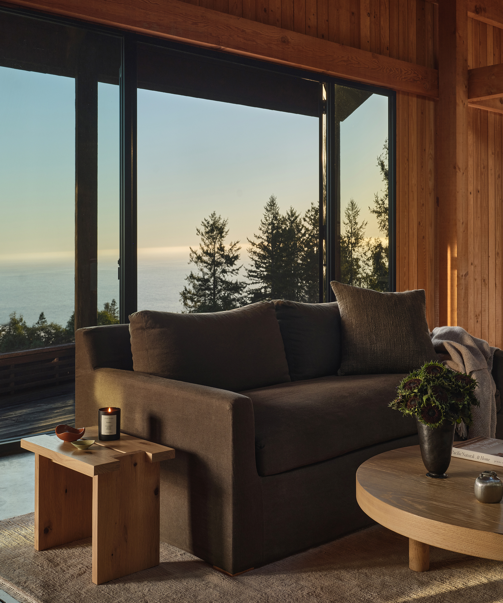 A cozy living room with a dark sofa, wooden tables, and floor-to-ceiling windows overlooking pine trees and the ocean at sunset. Warm natural light fills the wood-paneled space, perfect for spring decorating inspiration.