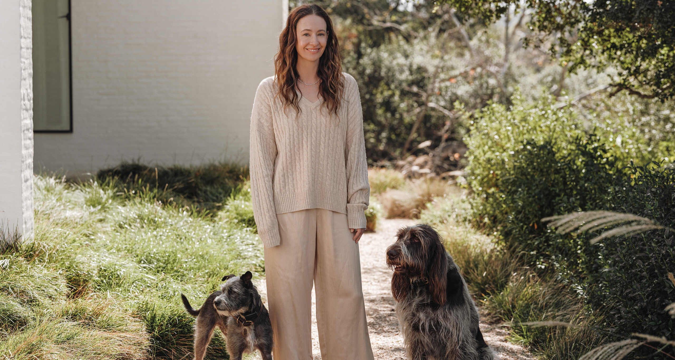 A woman in a beige sweater and wide-leg pants showcases her spring style outdoors on a garden path, smiling with two dogs—one small and one large—by her side. Lush greenery and a white building form the backdrop.
