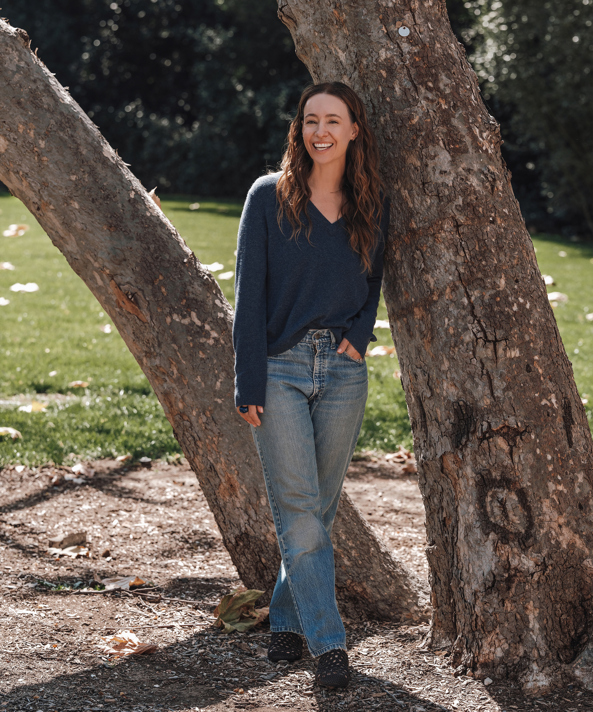 A woman with long brown hair, wearing a blue sweater and jeans in classic spring style, stands smiling and leaning against a tree in a sunny outdoor park.
