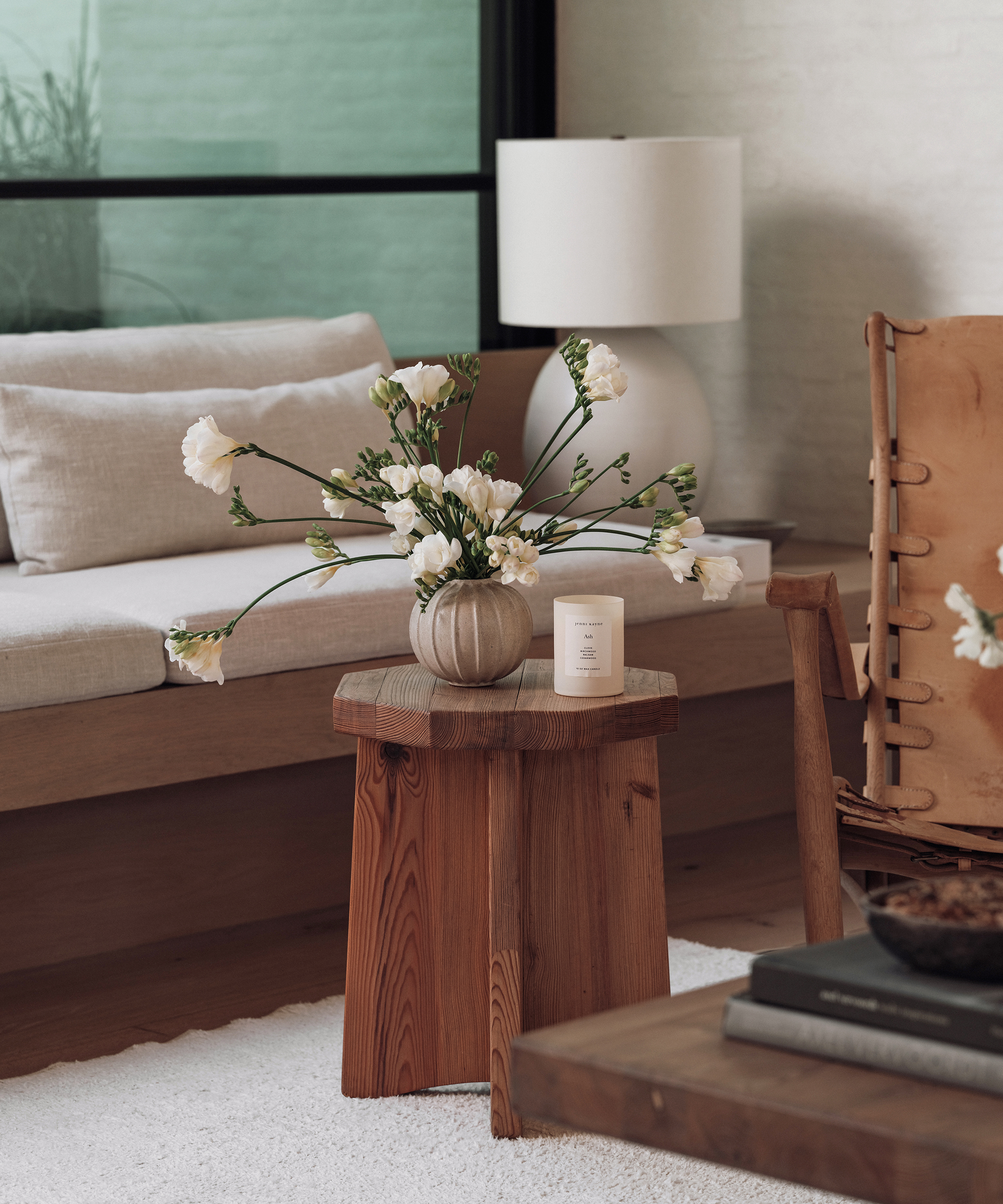 A small wooden table with a beige vase of white flowers and a white candle sits in a cozy, neutral-toned living room, adding a touch of spring decor. Nearby are a modern lamp, cushioned bench, and leather chair.