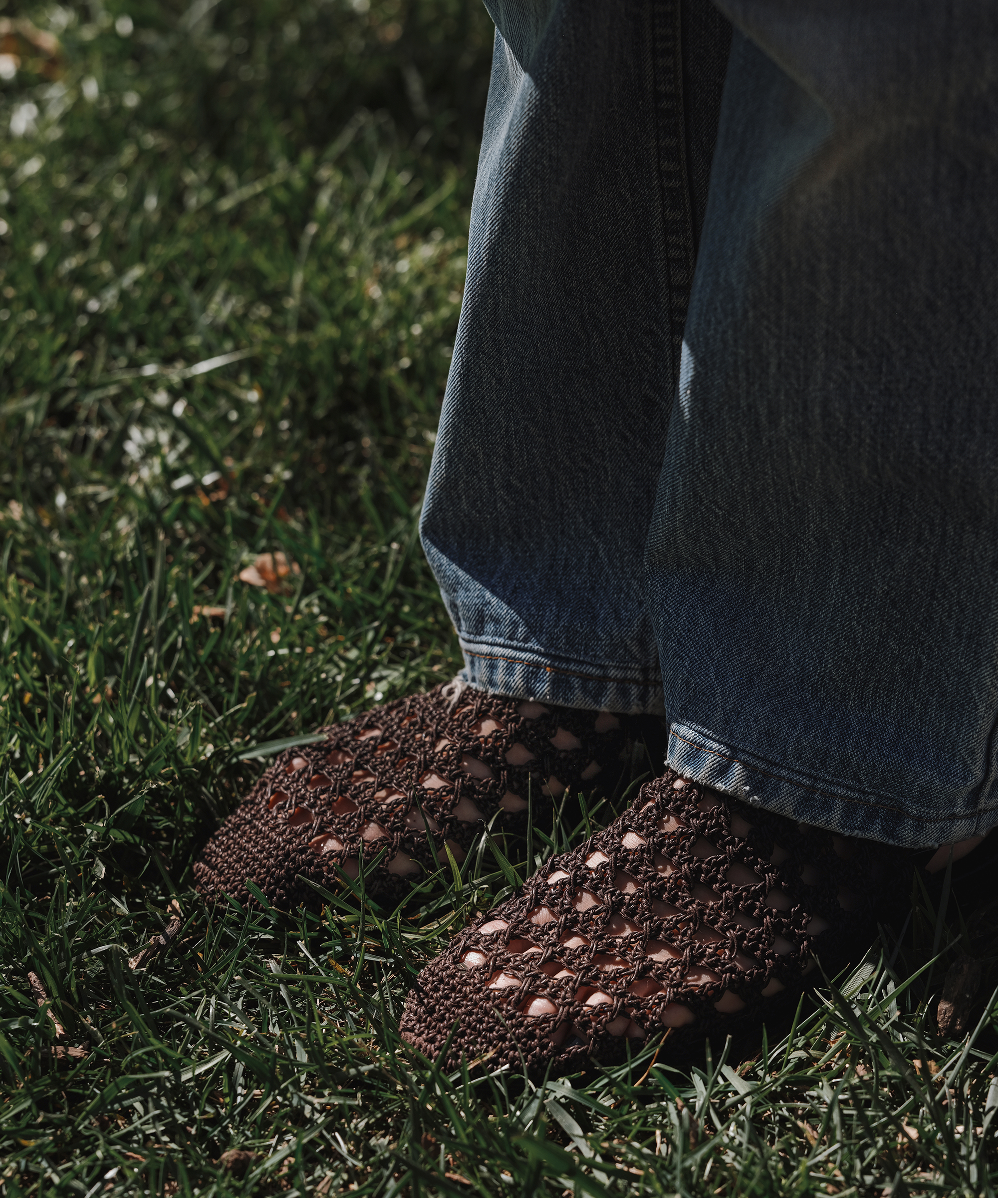 Person wearing blue jeans and brown woven shoes stands on green grass in sunlight, showcasing fresh spring style.
