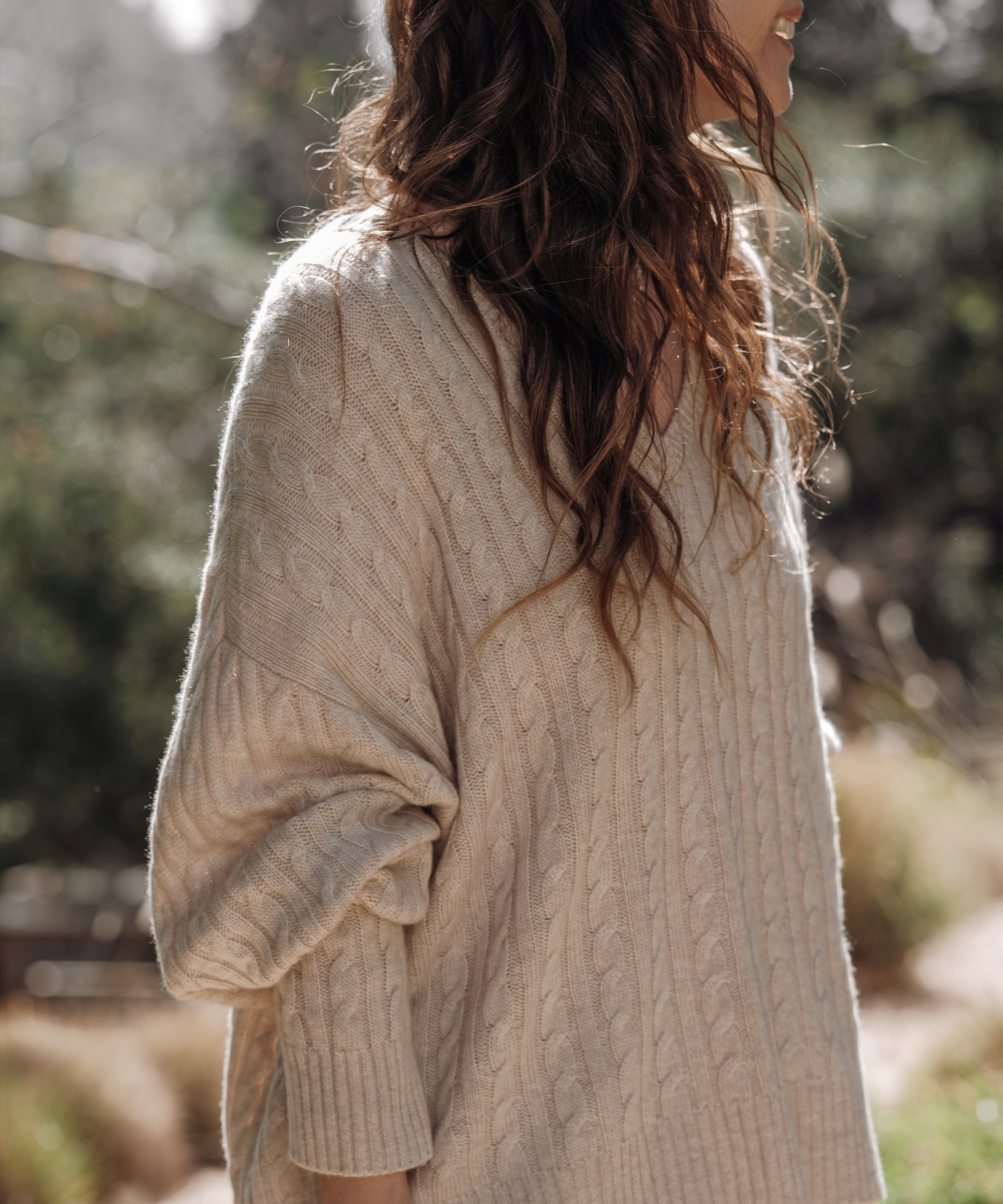 A woman with long wavy brown hair wearing a loose cream-colored cable knit sweater stands outdoors, embracing effortless spring style amid sunlight and greenery. Her face is turned away, partially out of view.