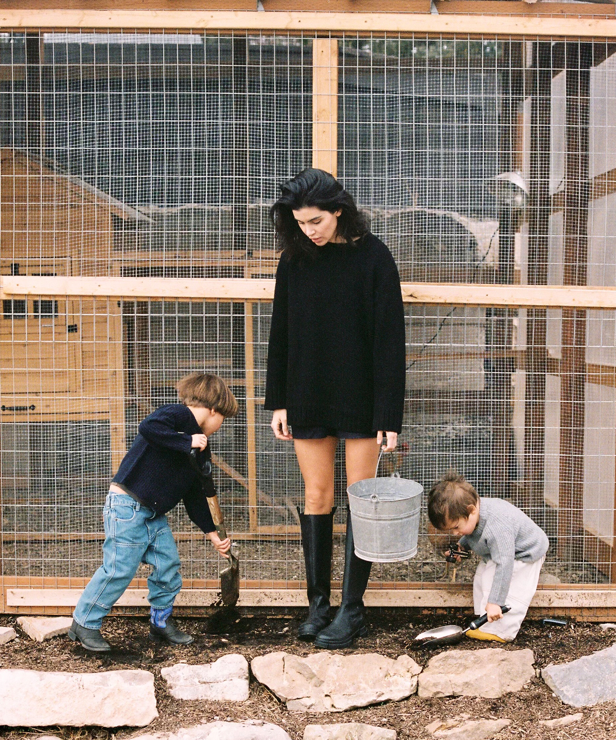 sara in a black cashmere sweater with a tin bucket and her two kids helping by the barn