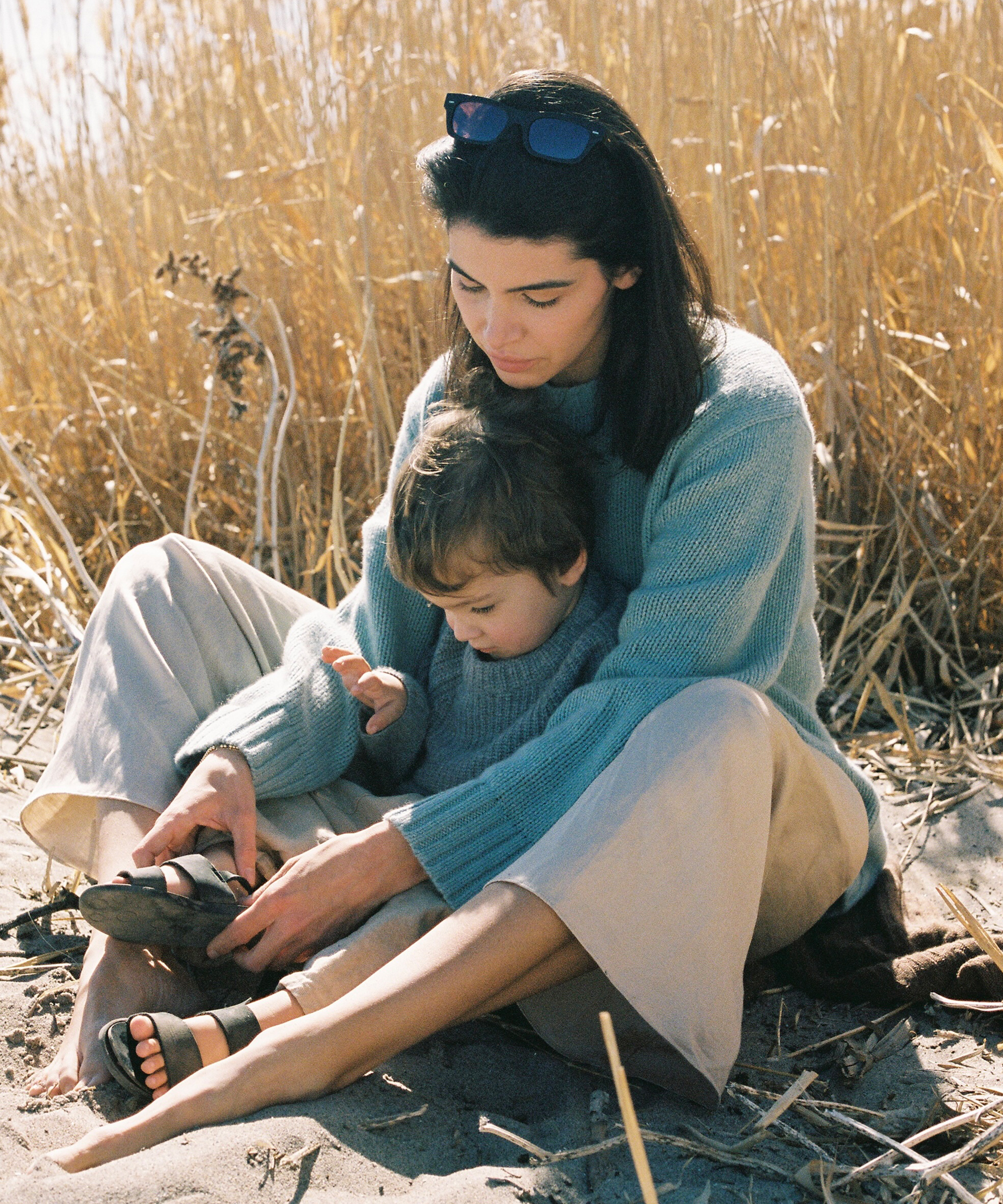sara wearing a blue cashmere sweater and oatmeal linen pants sitting on the beach with her son in a cashmere fisherman sweater