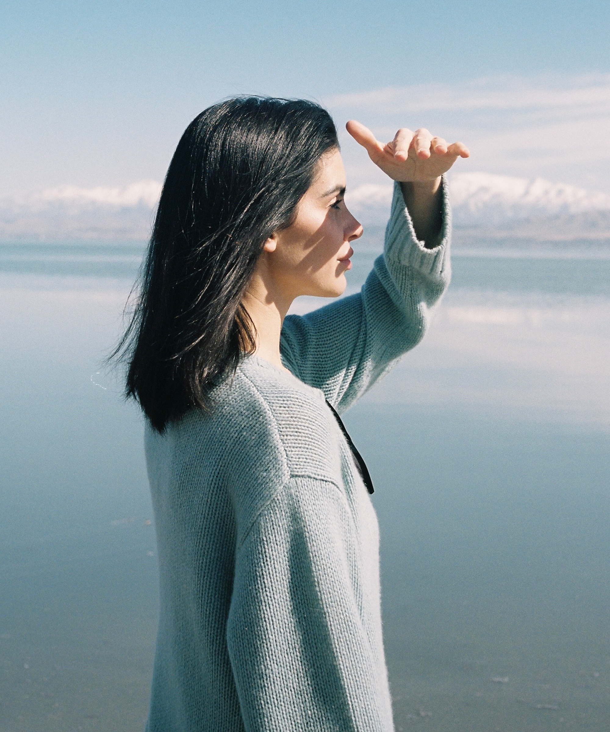 Sara Covey, a woman with dark hair in a light blue sweater, stands by a calm lake, shielding her eyes from the sun. Snow-capped mountains and a blue sky stretch out in the background.