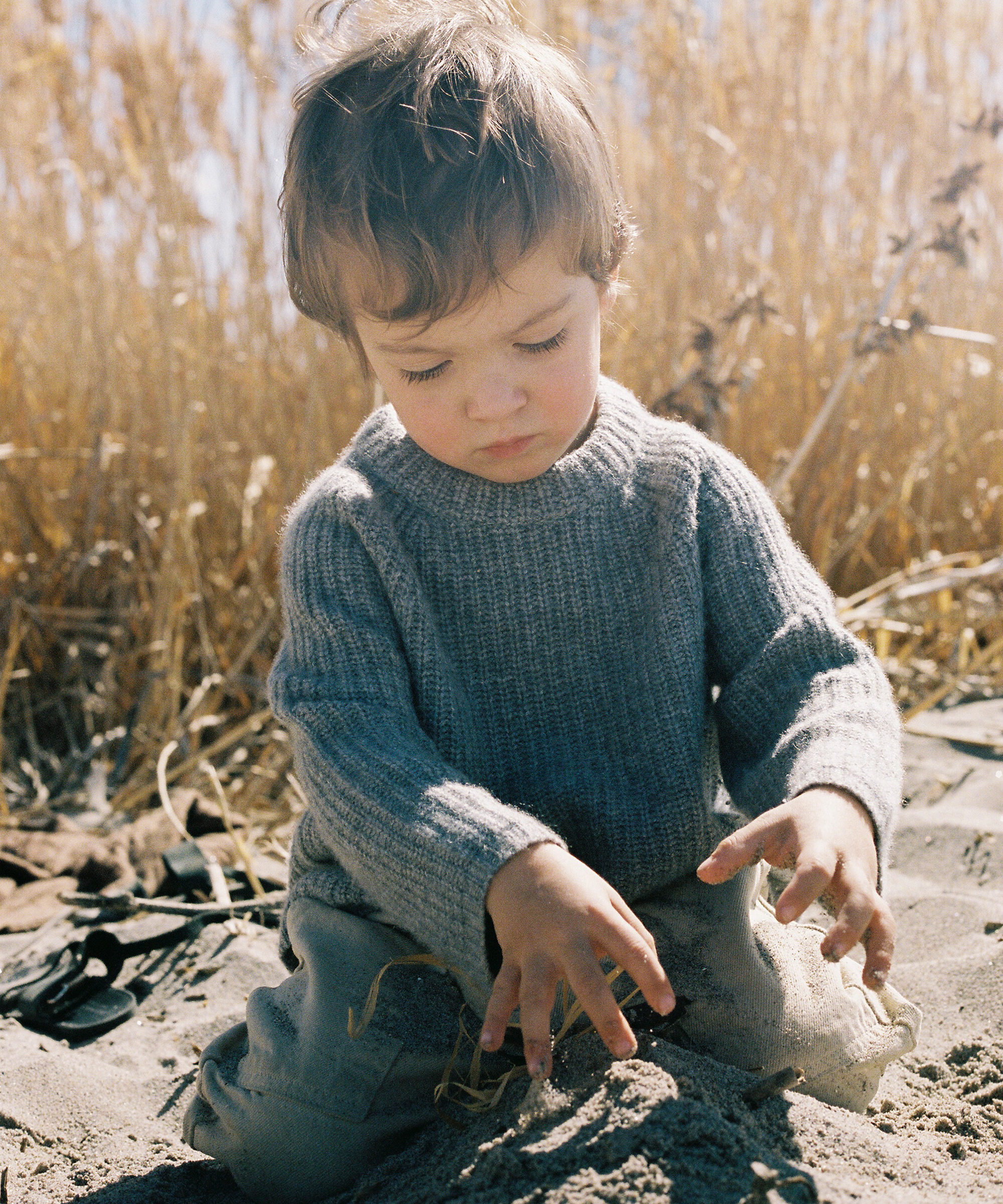 a little boy in a grey cashmere fisherman sweater playing with sand on the beach