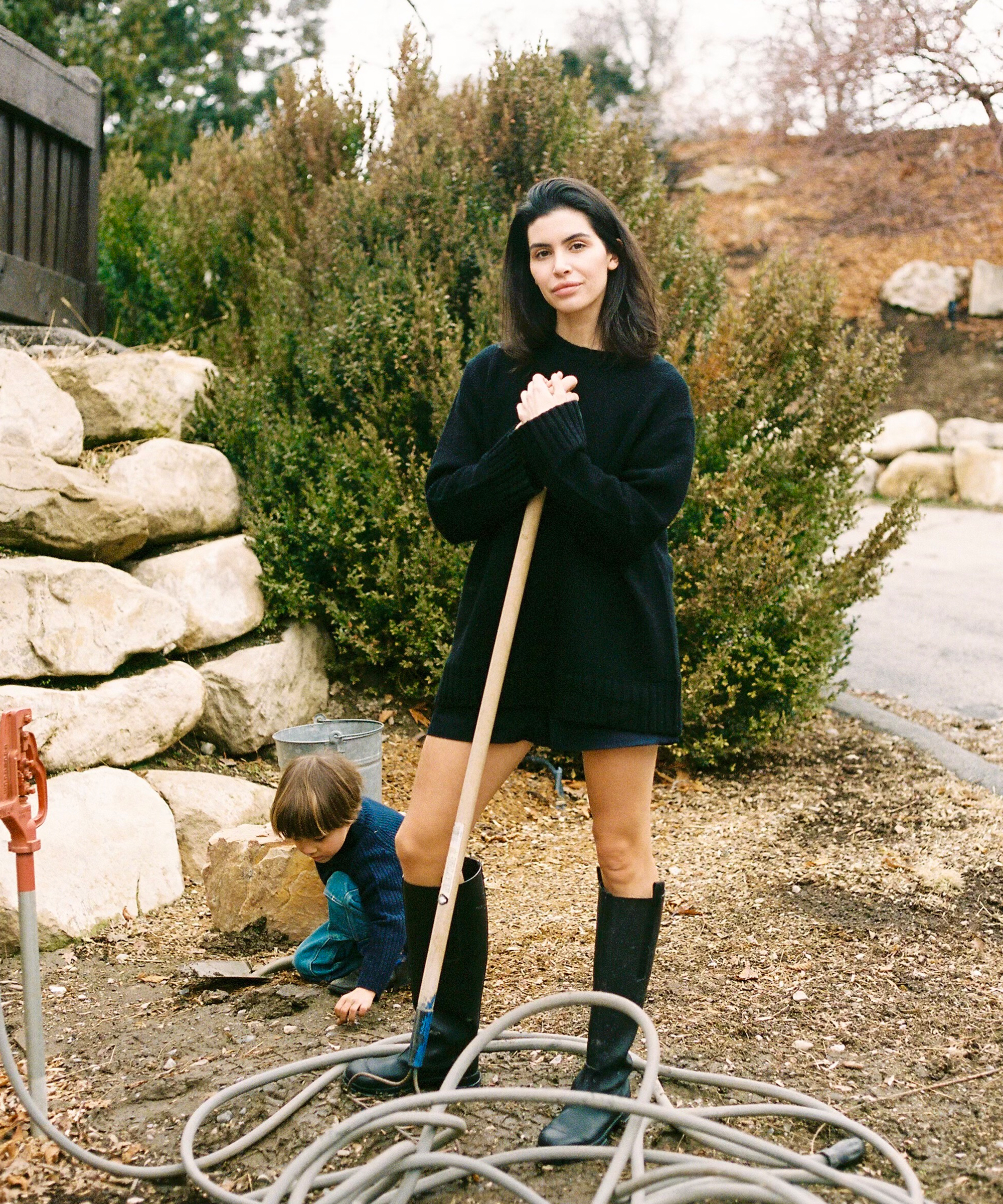 sara wearing a black cashmere sweater and rain boots shoveling in the yard
