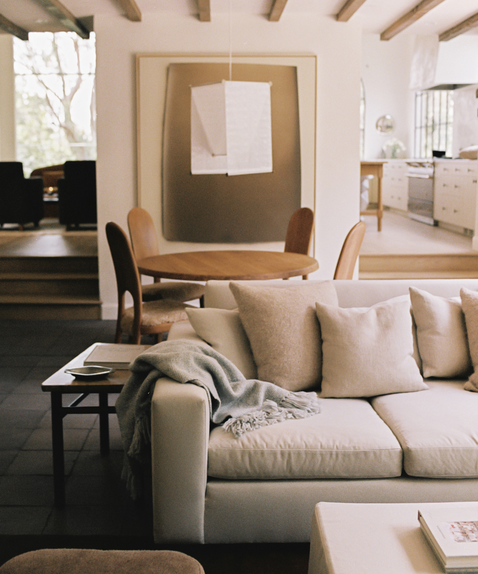 A cozy living room with a cream sofa, soft cushions, and a light blanket. Behind it is a round wooden dining table with chairs, large neutral wall art, and a modern pendant lamp. The space looks bright and inviting.