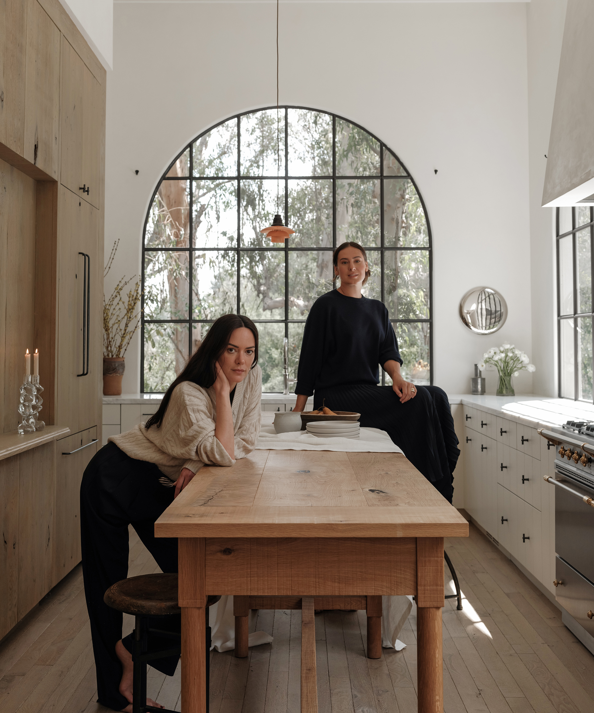 Two women pose in a modern, bright kitchen with large arched windows. One woman leans on a wooden table, while the other sits on the countertop. The space features light wood, white cabinets, and natural light.