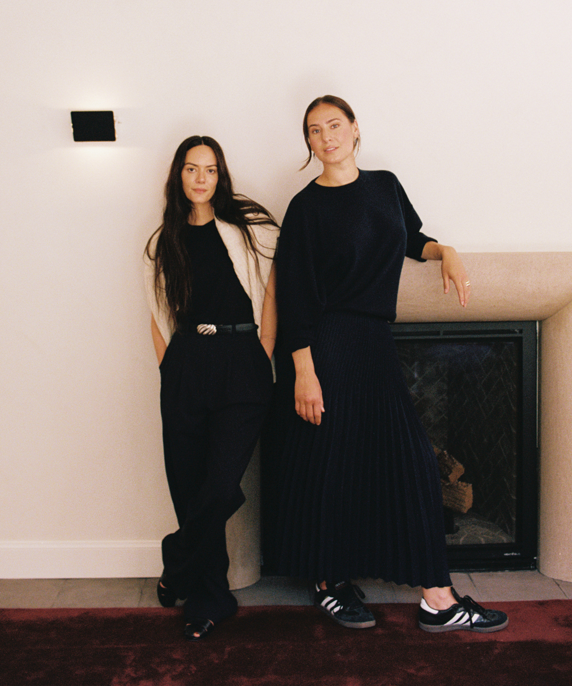 Two women stand indoors by a fireplace. One wears a black top, pleated skirt, and sneakers; the other wears a short-sleeve top, trousers, and sneakers. Both have long dark hair and neutral expressions.