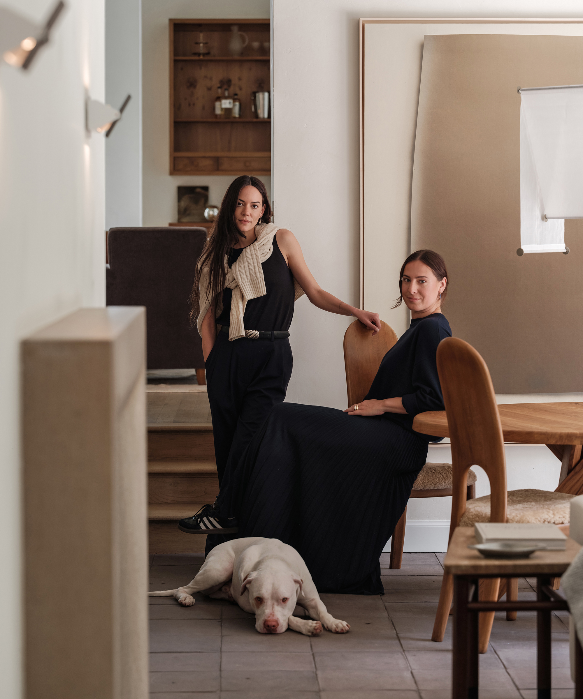 Two women with long brown hair pose in a modern, minimalist room. One stands with a sweater draped over her shoulders; the other sits at a wooden table. A white dog lies on the floor in front of them.