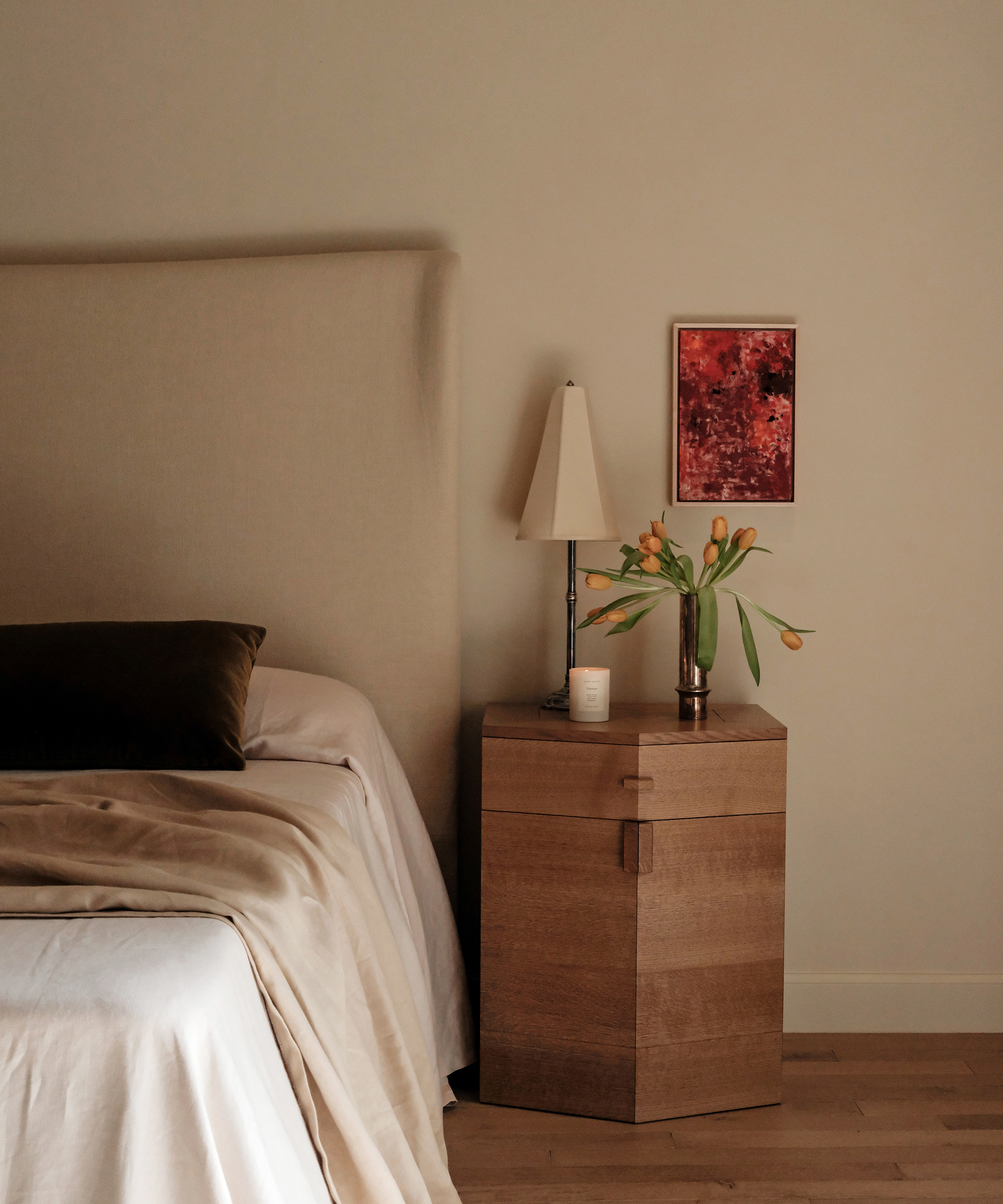 A minimal bedroom scene with a beige bed, dark pillow, wooden nightstand, lamp, candle, vase of yellow tulips, and a small red abstract painting on a plain wall. Warm, natural lighting gives a cozy feel.