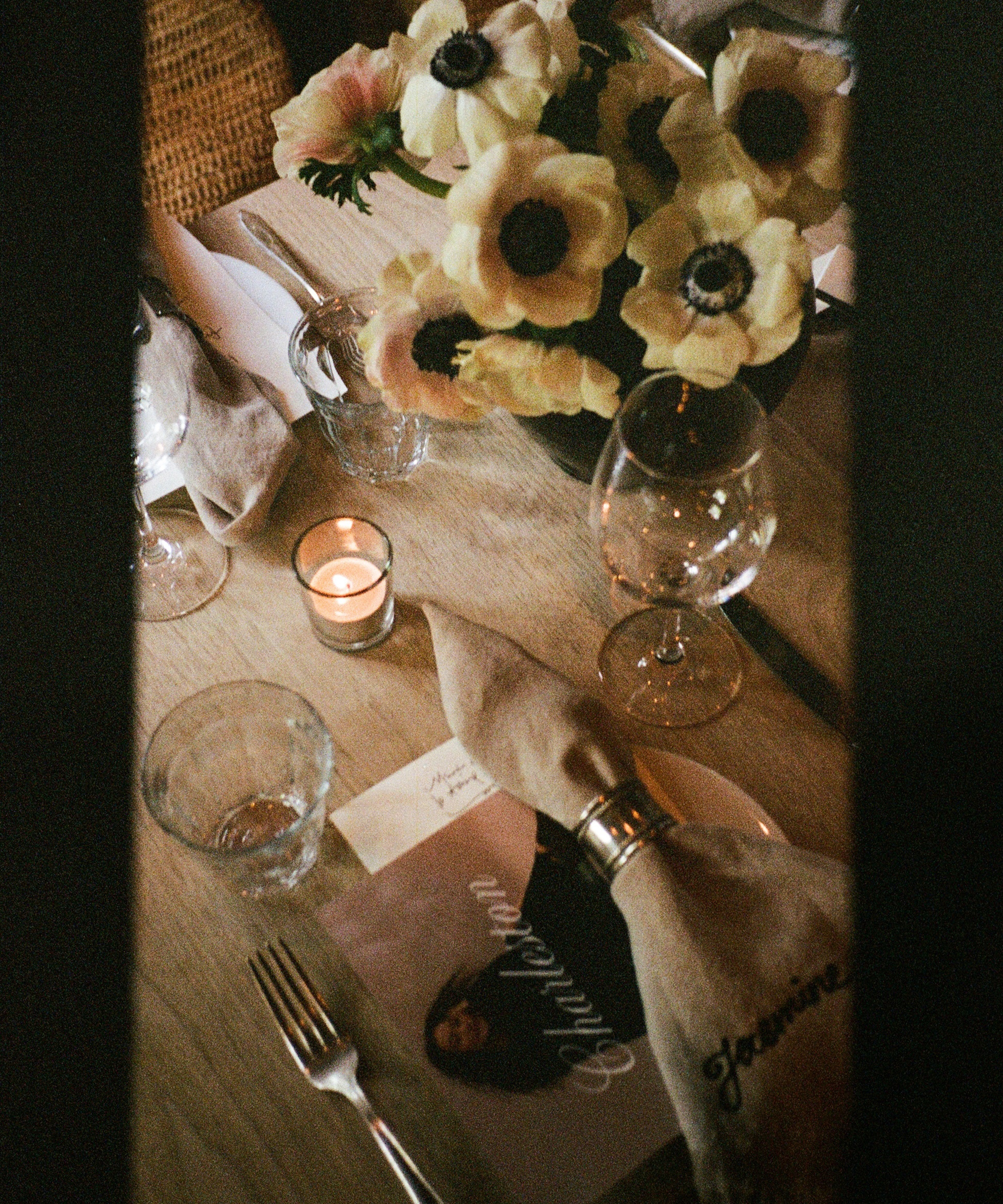 A warmly lit table set for a meal in Charleston, adorned with napkins, wine glasses, a candle, pale flowers, and a booklet featuring a portrait and the word Children.