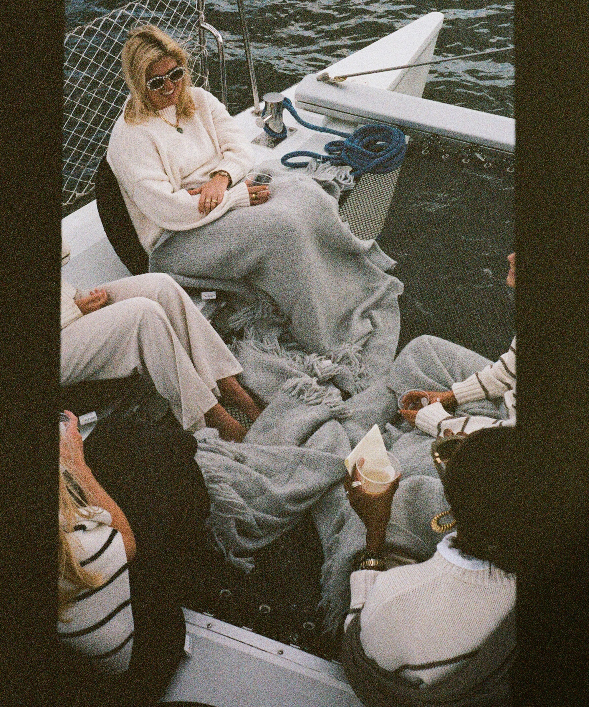 Four people sit on a boat near Charleston, wrapped in grey blankets and holding drinks. The group appears relaxed, dressed in light sweaters and pants. The sea is visible in the background, suggesting a cool, calm day.