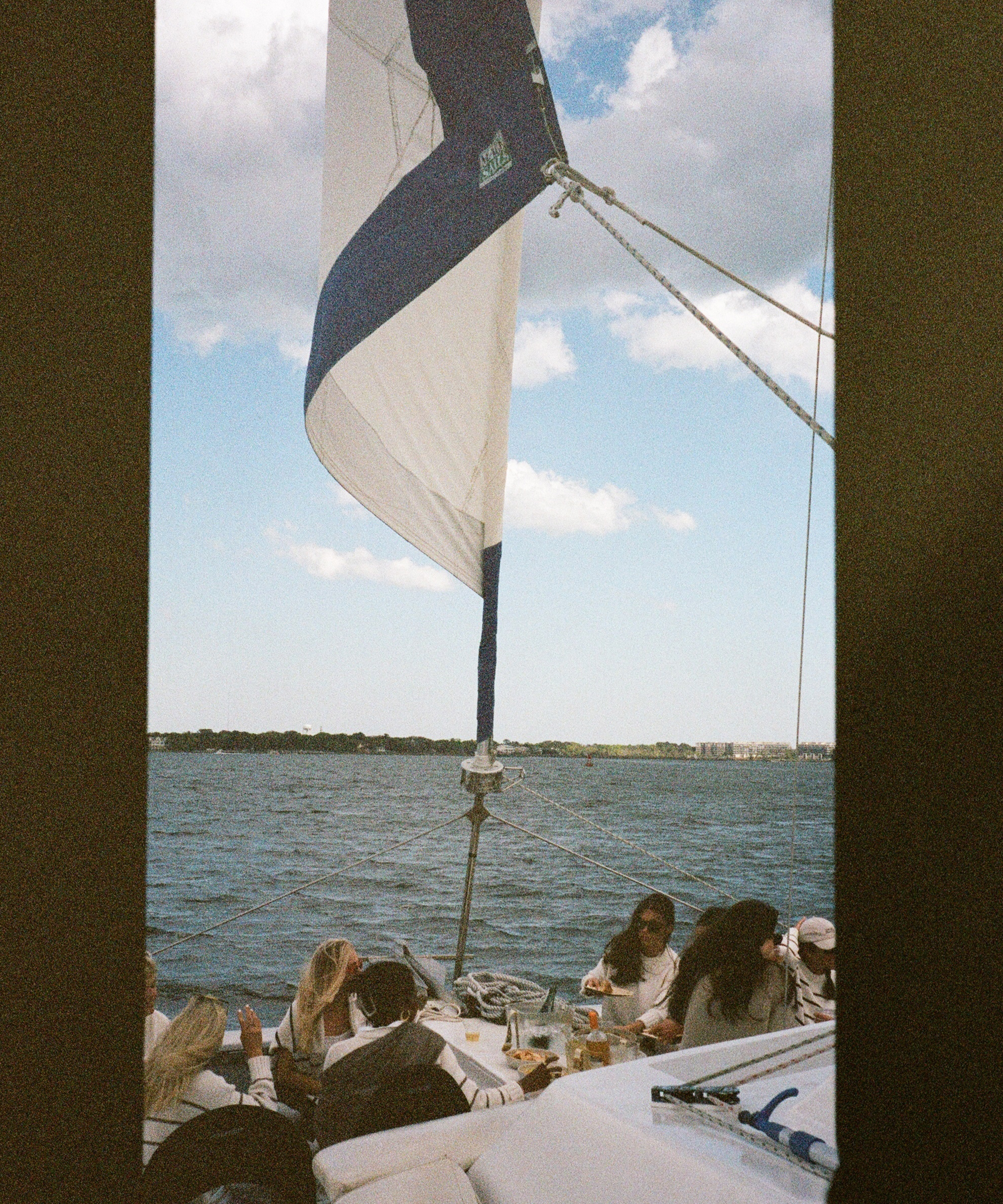 A group of people relax and socialize on a sailboat near Charleston under a partly cloudy sky, with a large sail raised and water and the distant shoreline visible in the background.